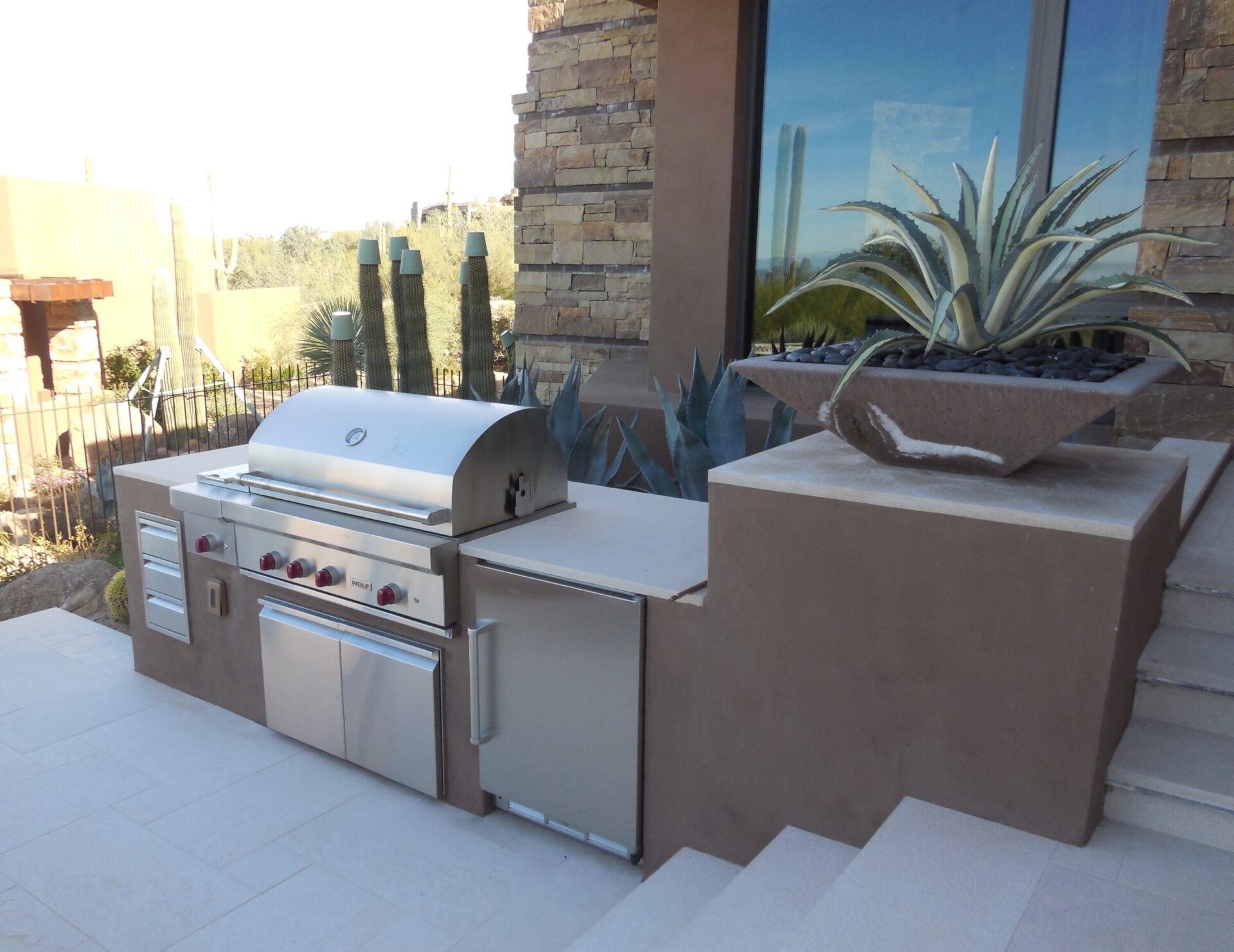 Outdoor modern grill setup beside a desert-themed garden. Features large agave plant, cacti, and contemporary architecture with stone accents.