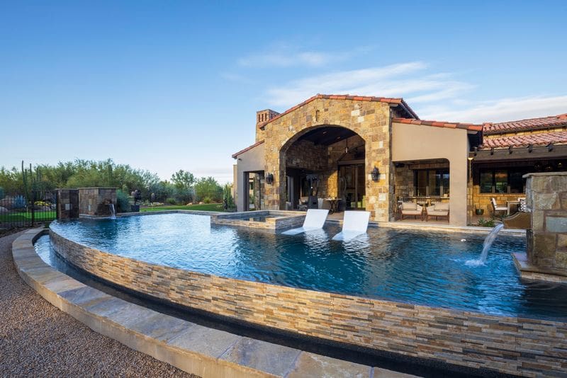 Modern luxury home with stone exterior, infinity pool, and sun loungers under a clear sky. Verdant landscape surrounds the serene, stylish residence.