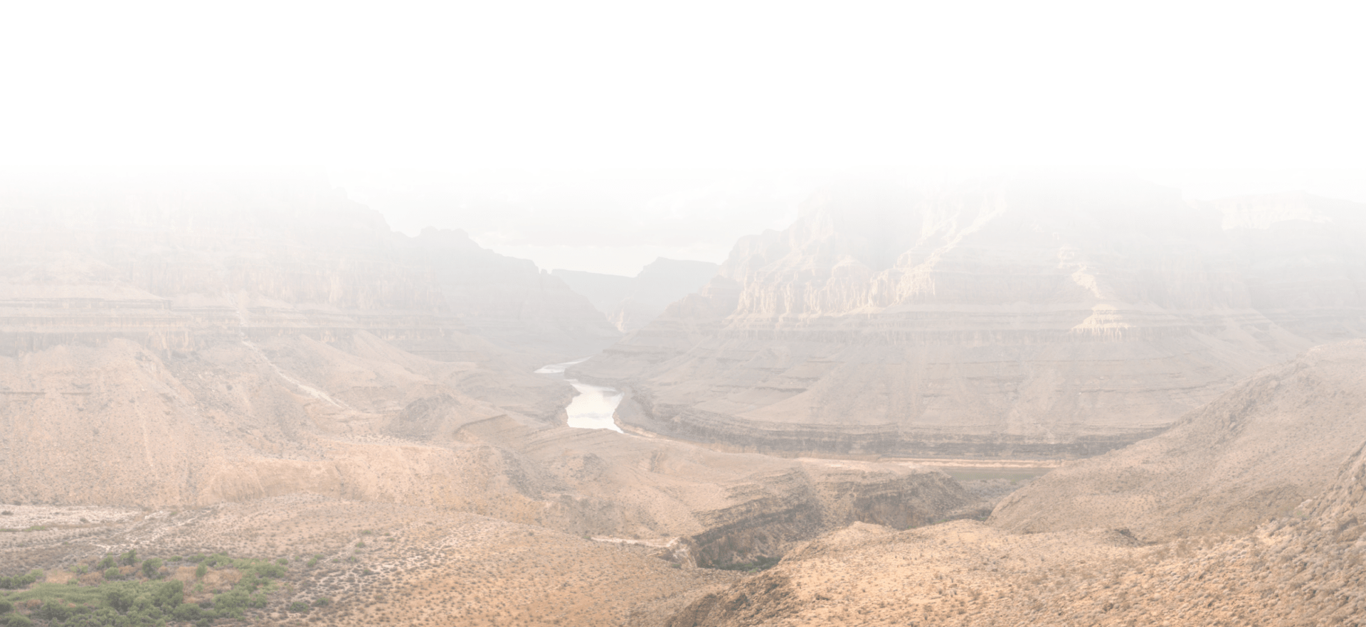 Misty view of the Grand Canyon, showcasing rugged cliffs and a winding river through the arid landscape, beneath a foggy sky.