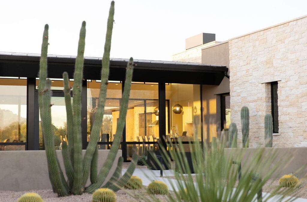 Modern house with large windows surrounded by desert landscape, including cacti and stone elements, under warm lighting. Interior visible through glass panes.