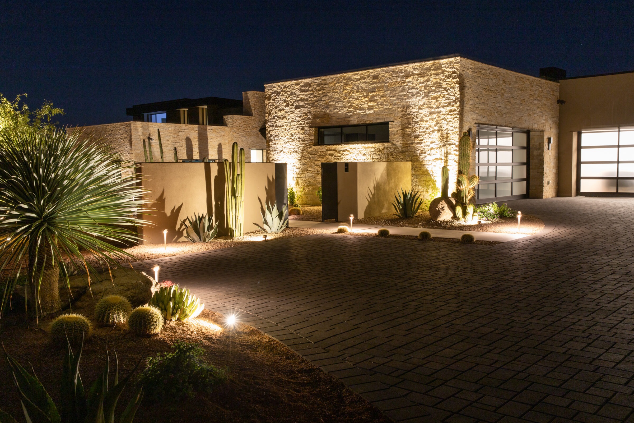 Modern desert home with illuminated stone facade, cacti landscaping, and a paved driveway, viewed at night with soft outdoor lighting accents.