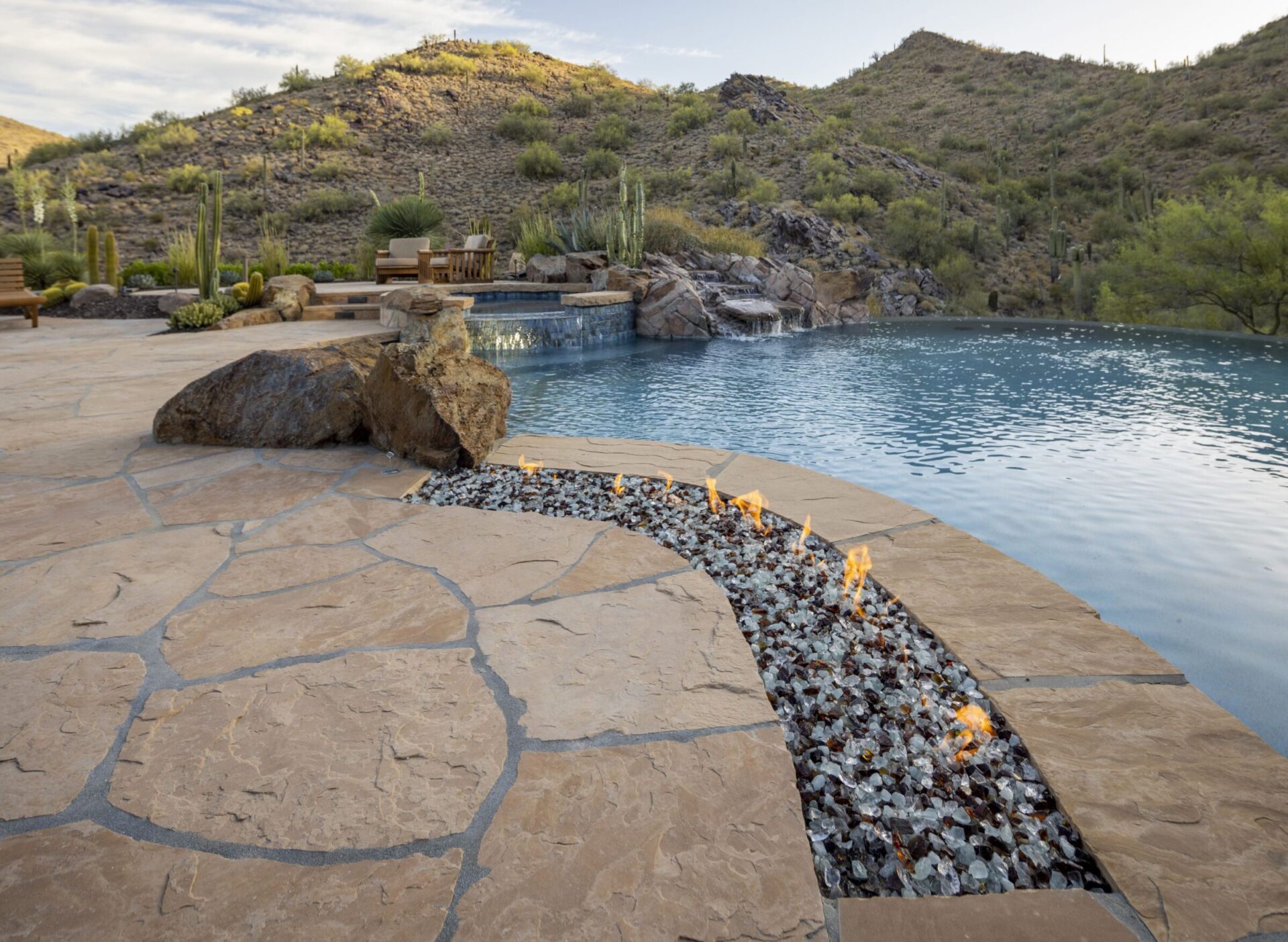 A serene desert landscape features an infinity pool, stone patio, fire pit, and distant mountains under a partly cloudy sky.