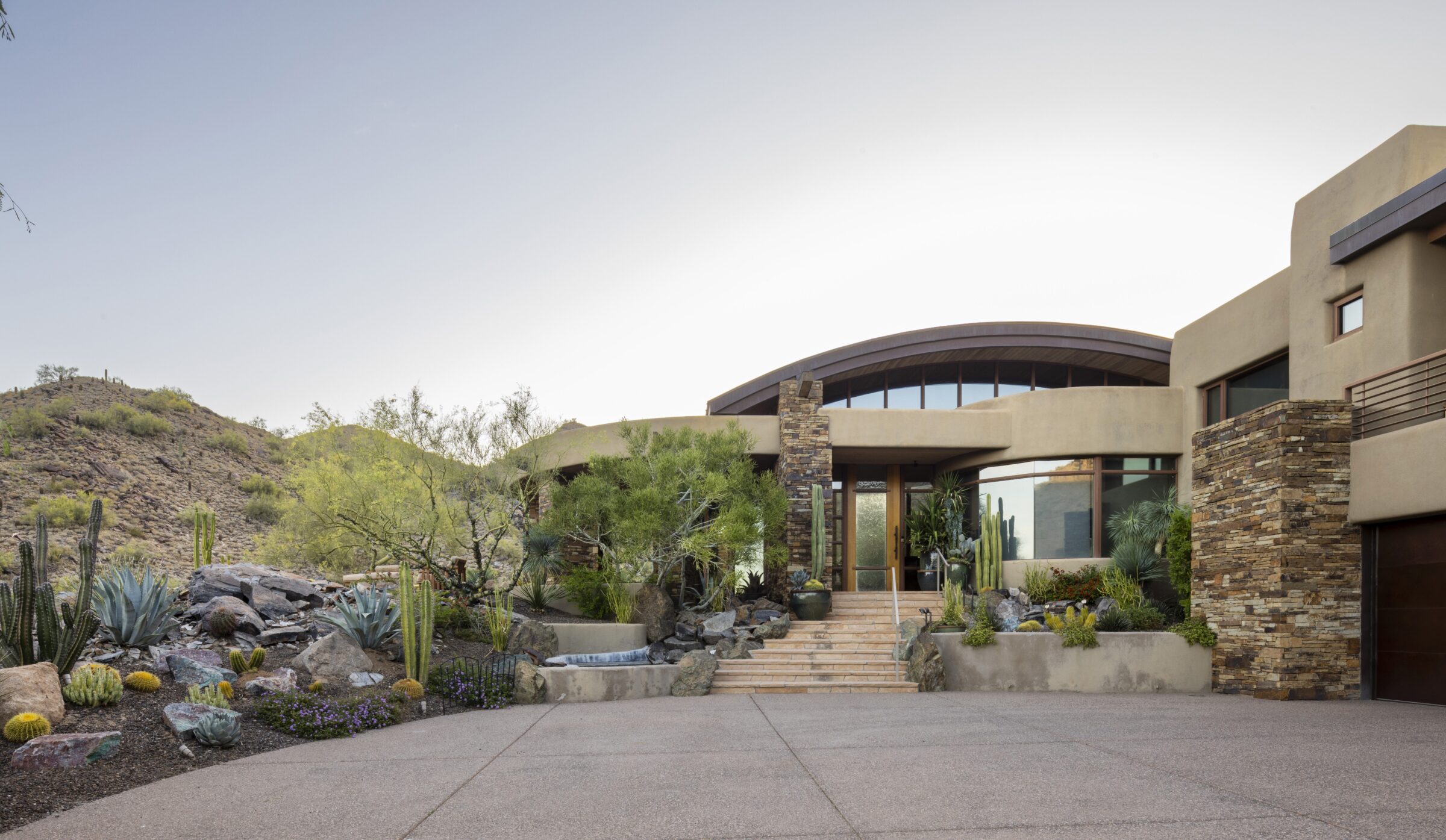 Modern house with desert landscaping, featuring cacti and stone elements. Surrounded by rocky hills under a clear sky. No people present.