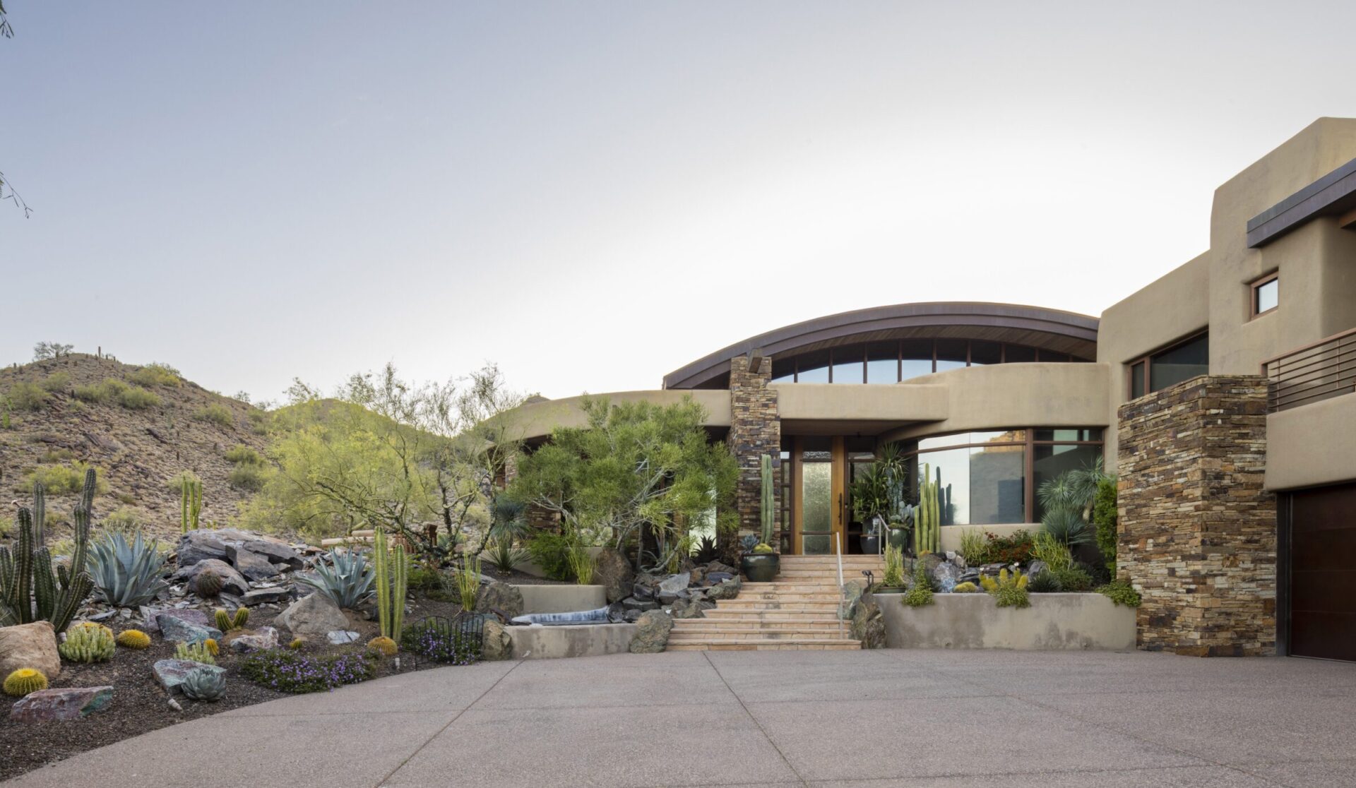 Modern house with desert landscaping, featuring cacti and rocks, set against a backdrop of rugged hills. Stone and stucco exterior design.