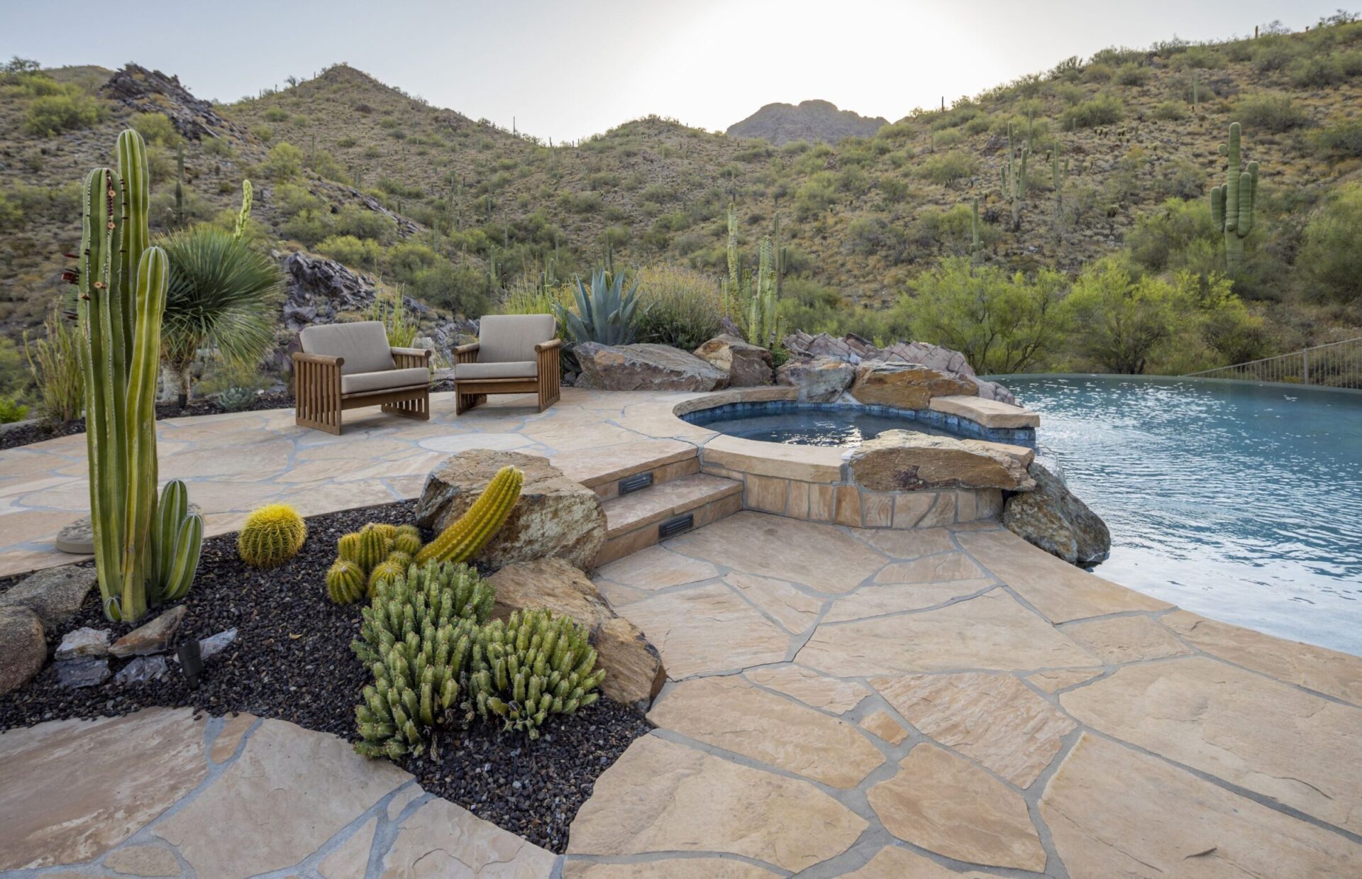 Desert landscape with cacti, two chairs, and a small pool on a stone patio, surrounded by hills under a clear sky.