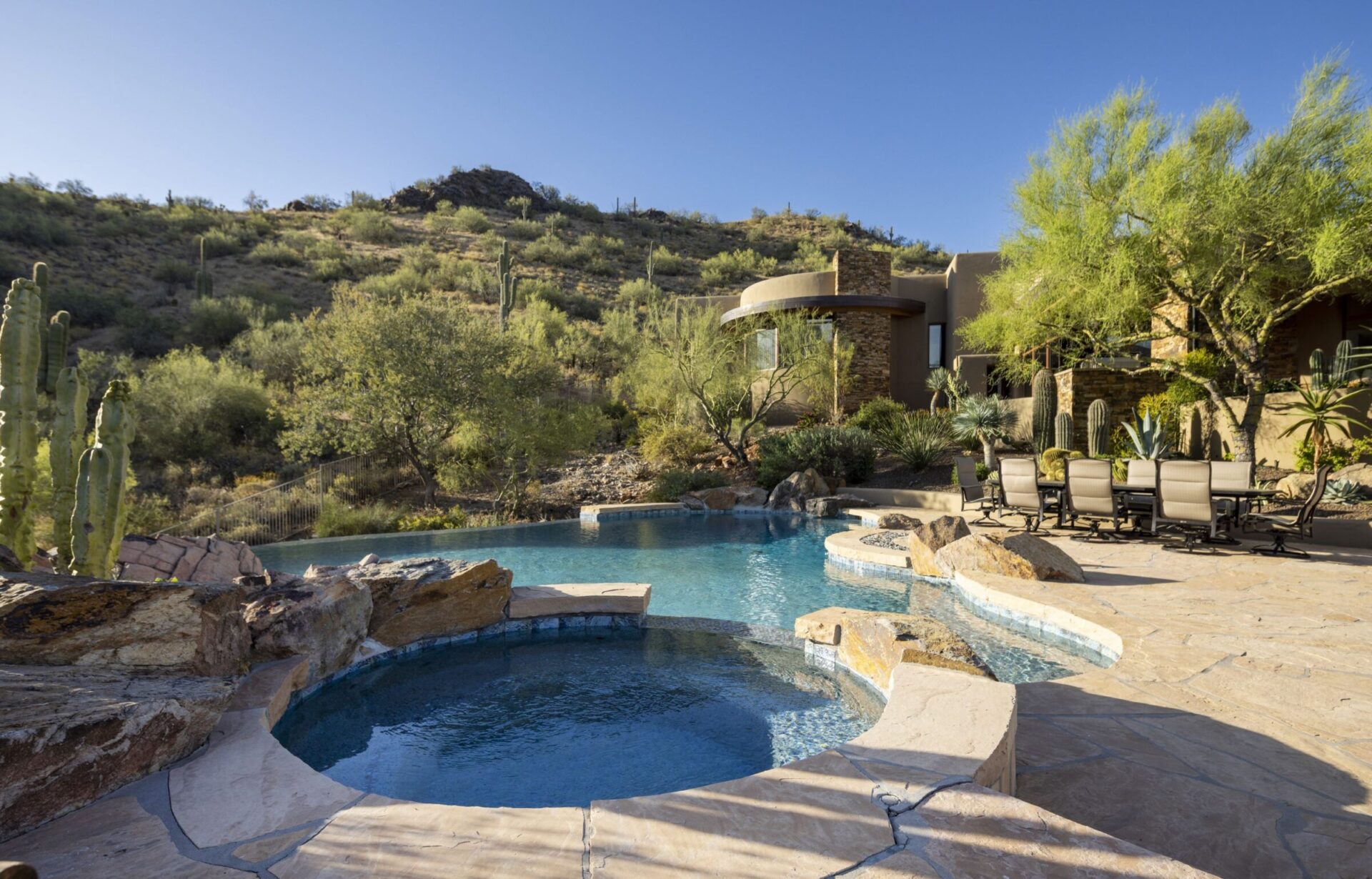 Desert home with pool and hot tub, surrounded by cacti and rocky landscape, under clear blue sky with hills in the background.