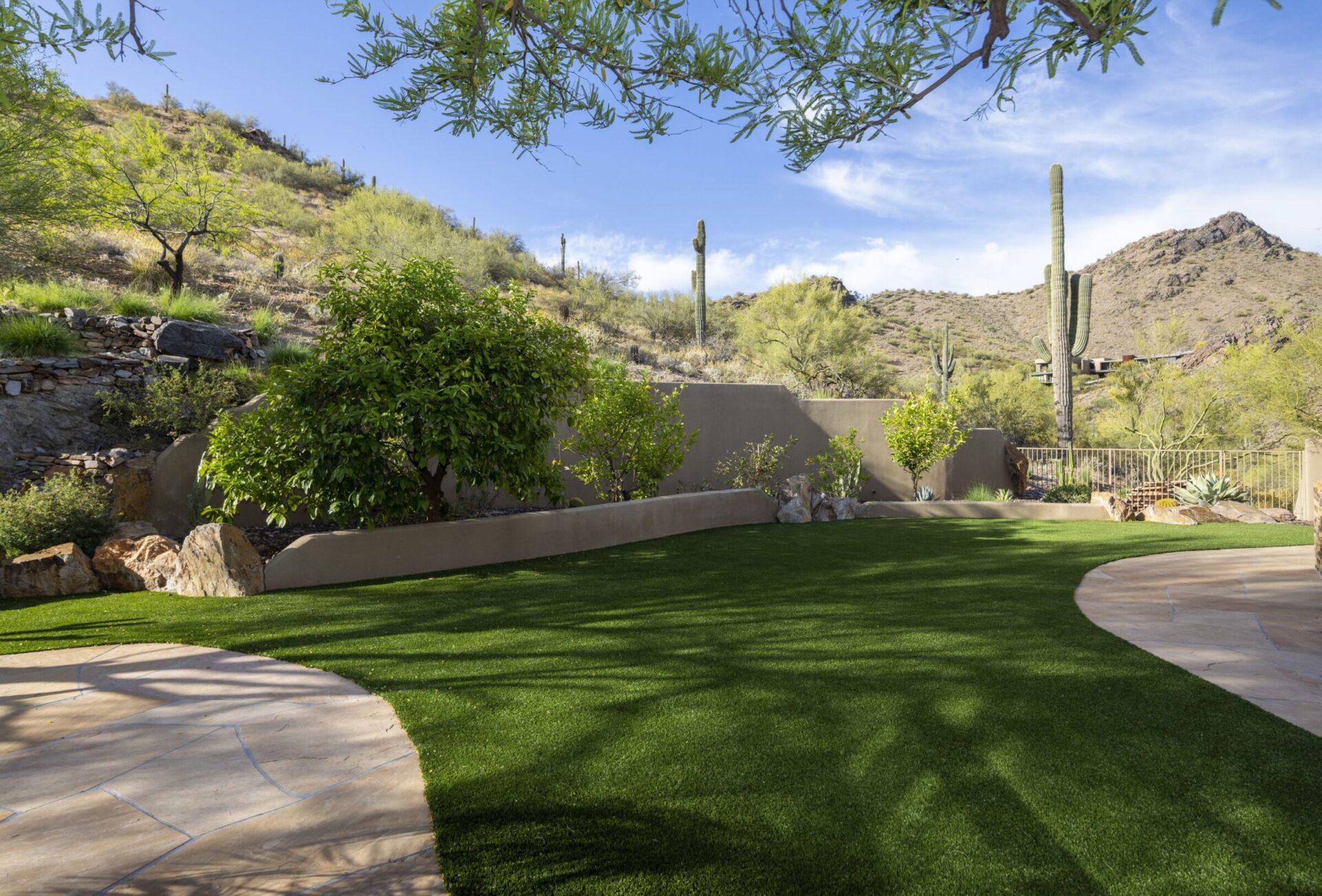 A landscaped garden with green grass, surrounded by desert hills and cacti under a bright blue sky, conveys a serene outdoor atmosphere.