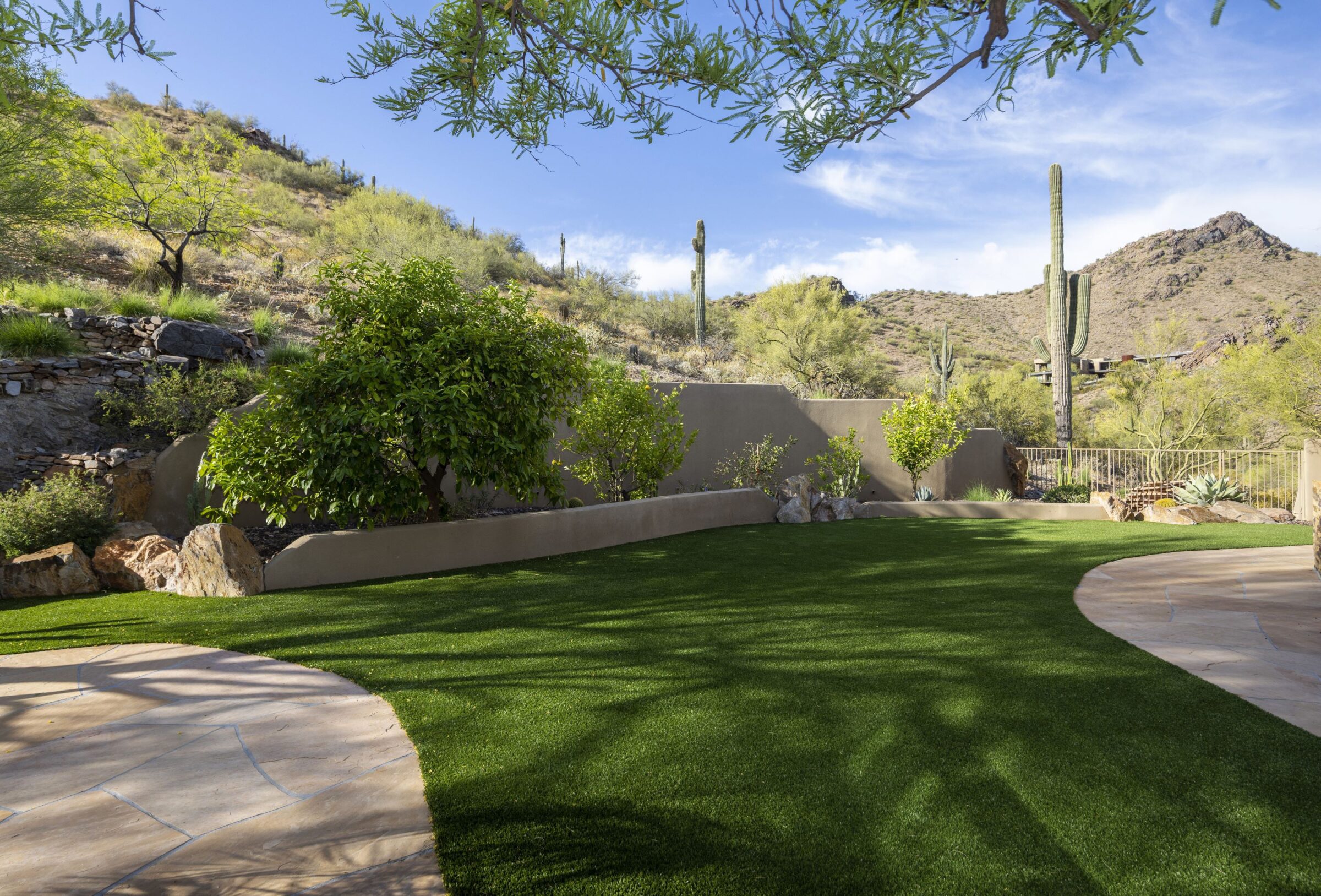 A landscaped garden with artificial grass, bordered by desert plants and cacti against a backdrop of rocky hills under a clear blue sky.