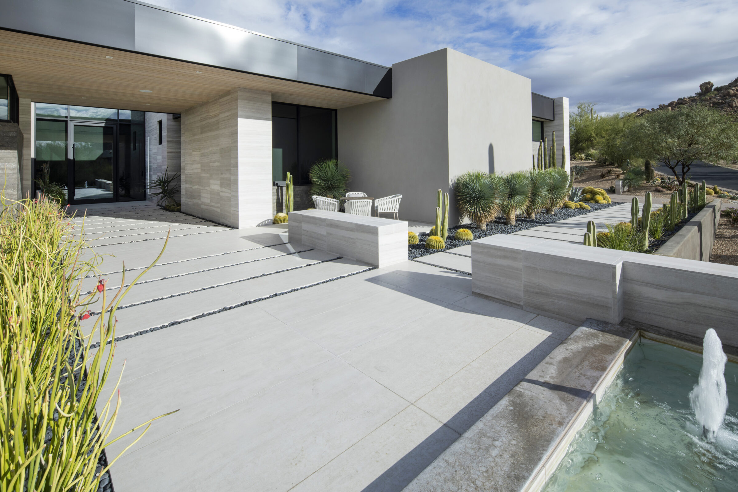 Modern house exterior with sleek lines, desert landscaping featuring cacti, and a small fountain in the foreground under a partly cloudy sky.