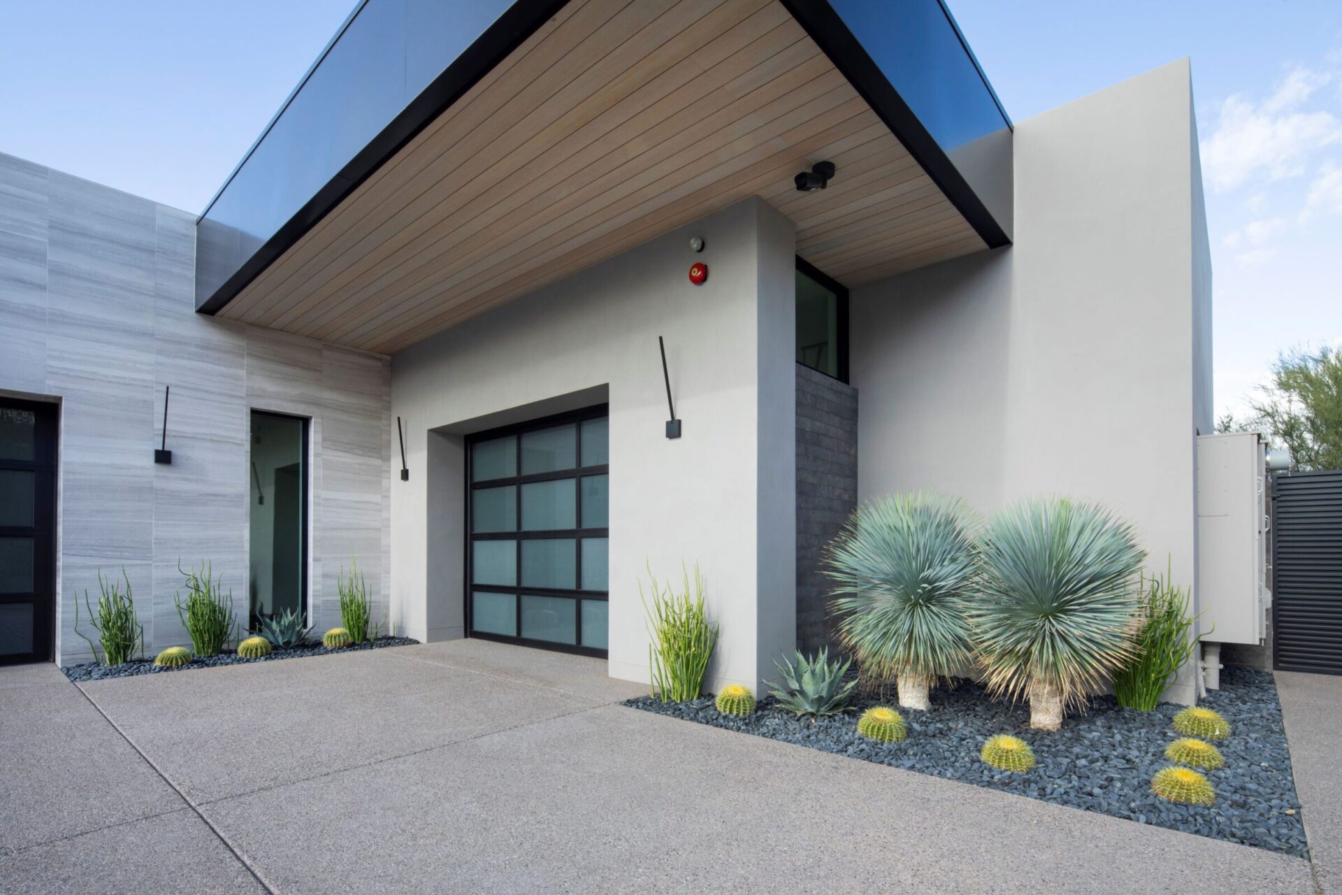 Modern house exterior with sleek design, minimalistic landscaping featuring cacti and succulents, glass garage door, concrete driveway, and contrasting materials. Contemporary architectural style.