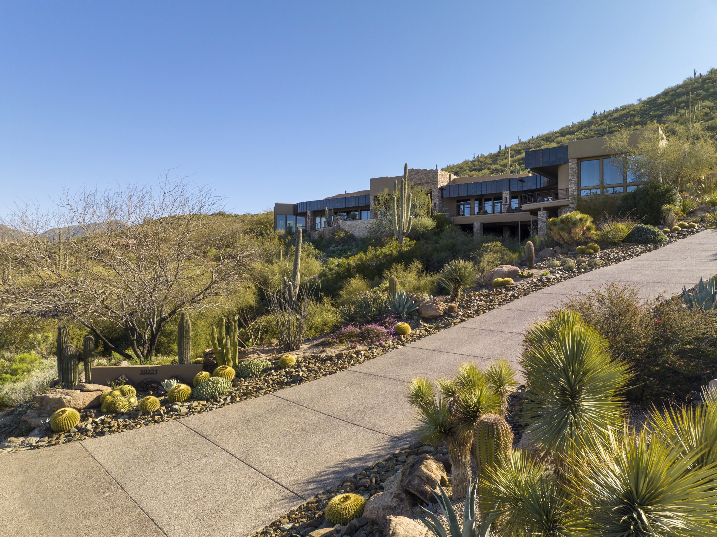 Modern house surrounded by desert landscape, featuring cacti and a sloping driveway leading towards the property under a clear blue sky.