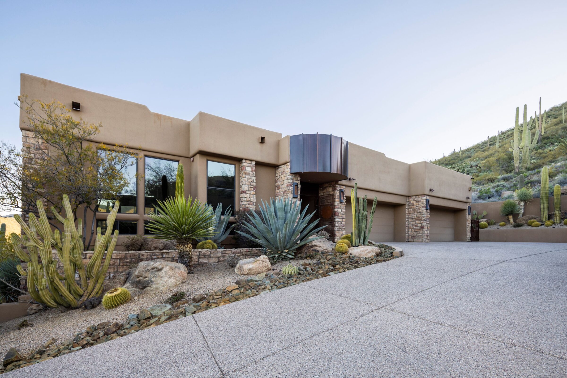 Modern desert-style house with stone and stucco facade, surrounded by various cacti and desert plants, set against a hill with more cacti.