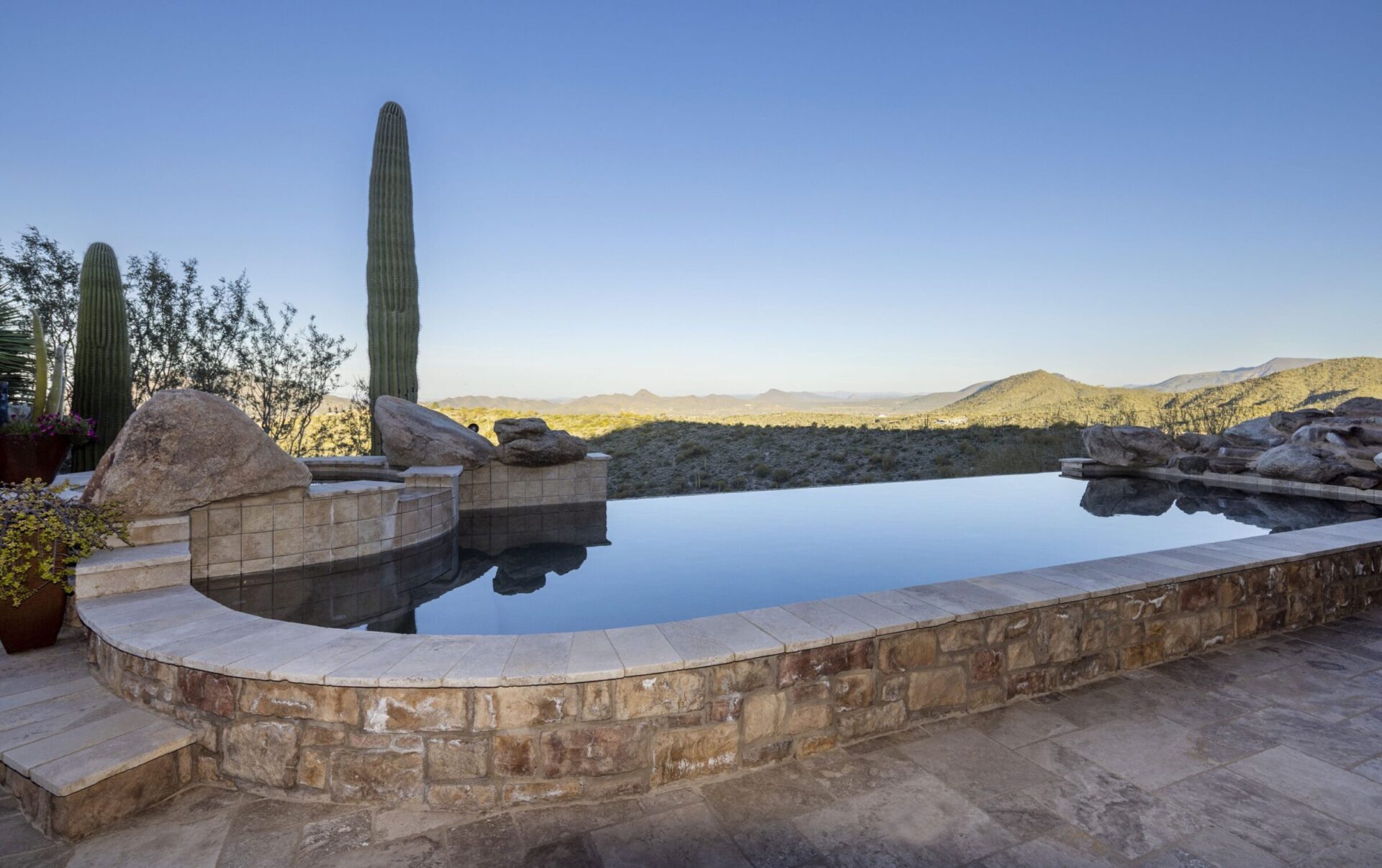 Infinity pool with stone tiles, surrounded by desert landscape, large cacti, and rocky formations under a clear blue sky. Peaceful and scenic.