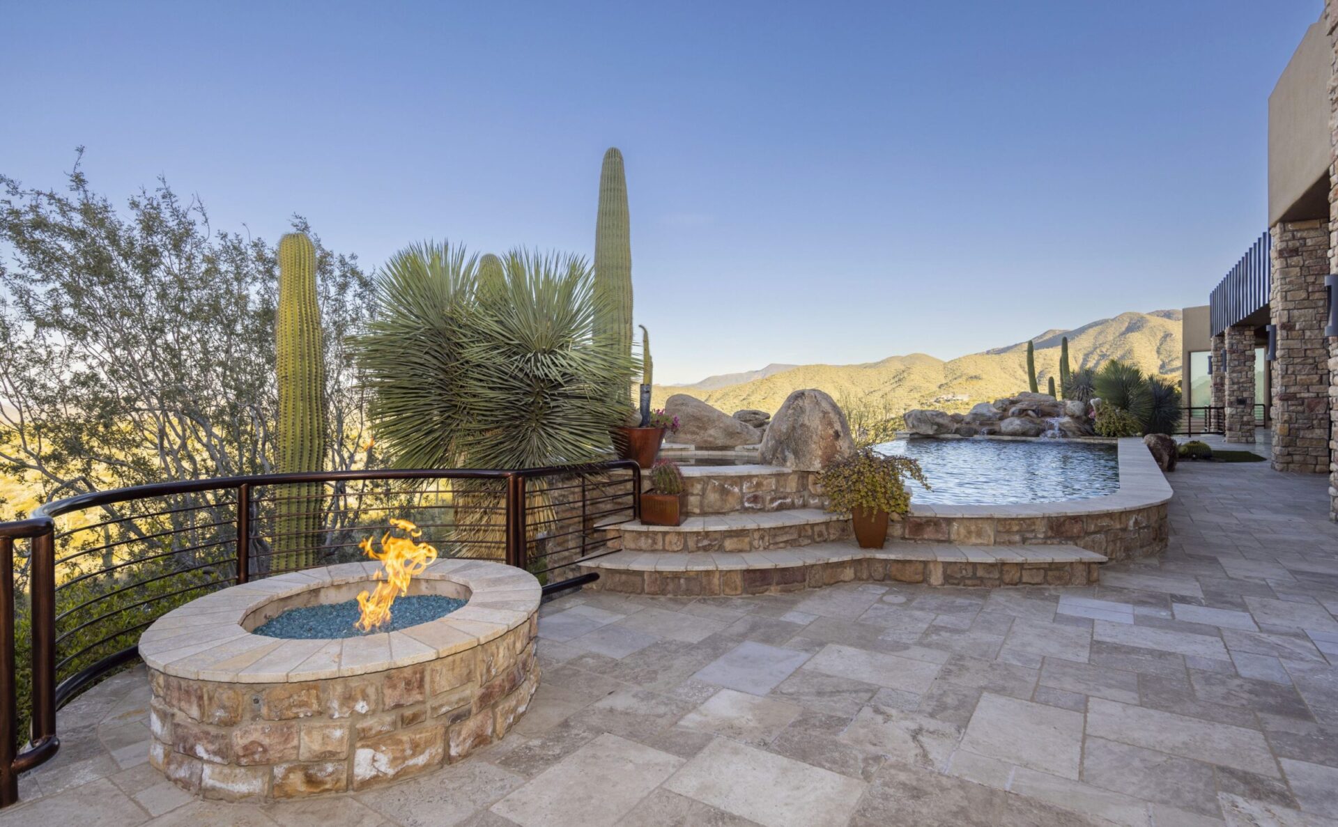 Desert landscape featuring a stone patio with a fire pit, pool, and cacti, overlooking distant mountains under a clear blue sky.