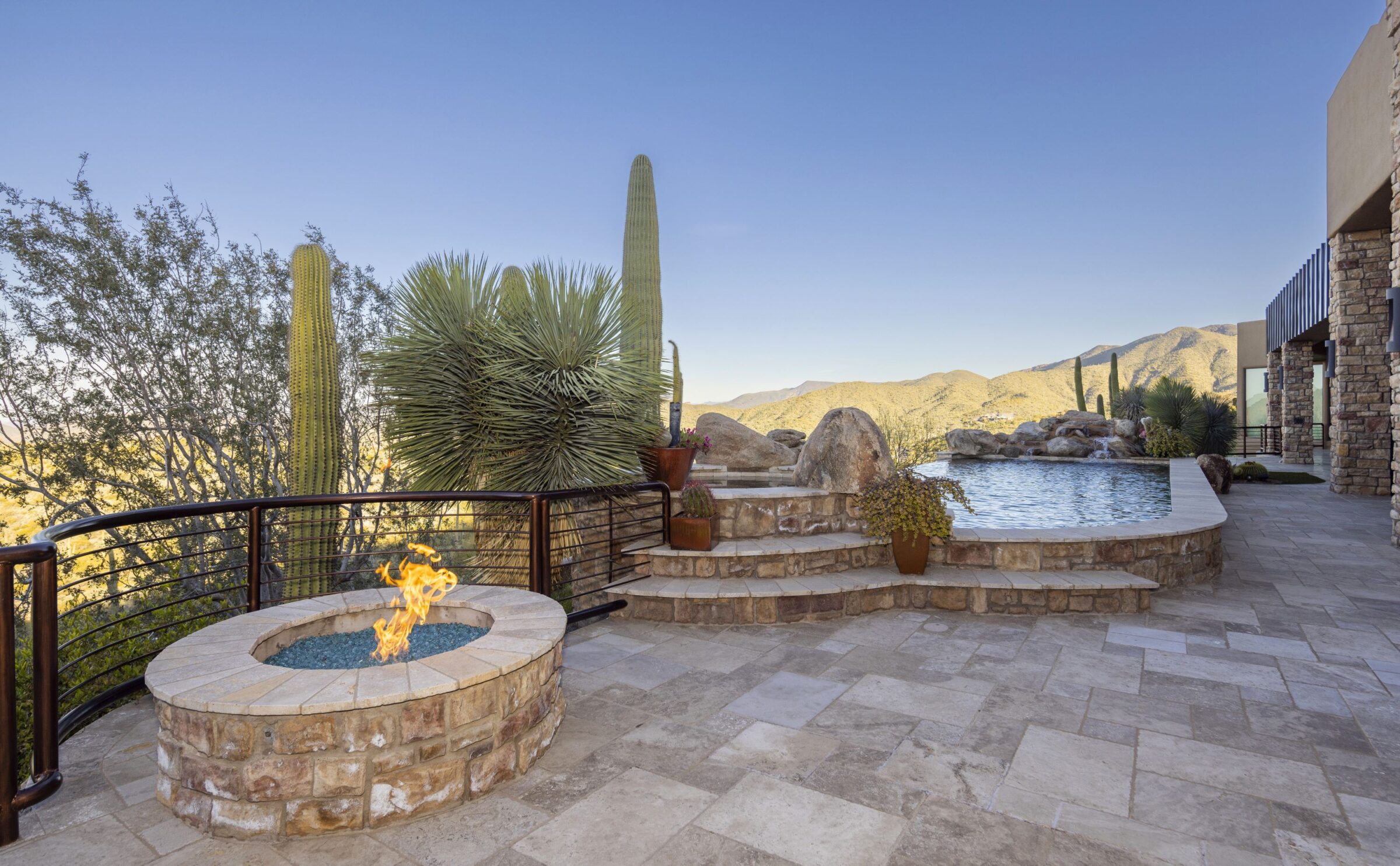 Stone patio with fire pit, pool, and desert landscape. Cacti and mountains in the background under a clear, blue sky.