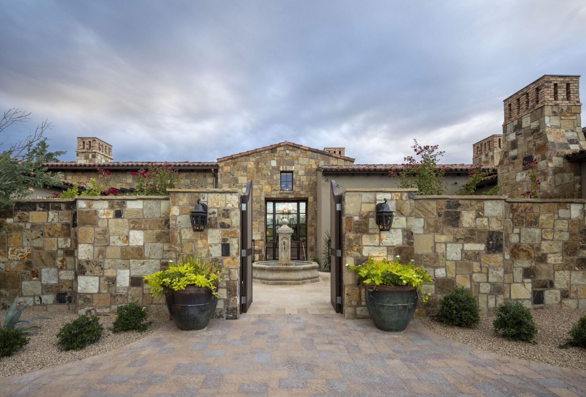 Stone courtyard entrance with a central fountain, flanked by potted plants and lanterns, leading to a rustic stone building.