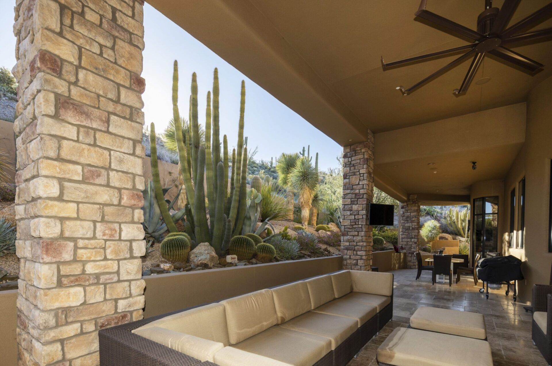 Covered patio with cushioned sofas and tables, surrounded by stone pillars and desert landscape featuring tall cacti and various desert plants.