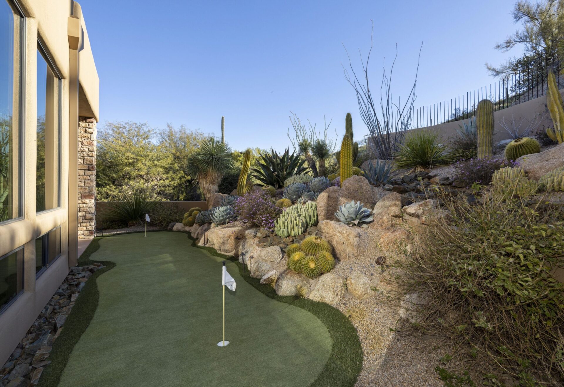 Modern house exterior with large windows, adjacent putting green, and cactus garden set against a clear blue sky. Desert landscaping enhances outdoor space.