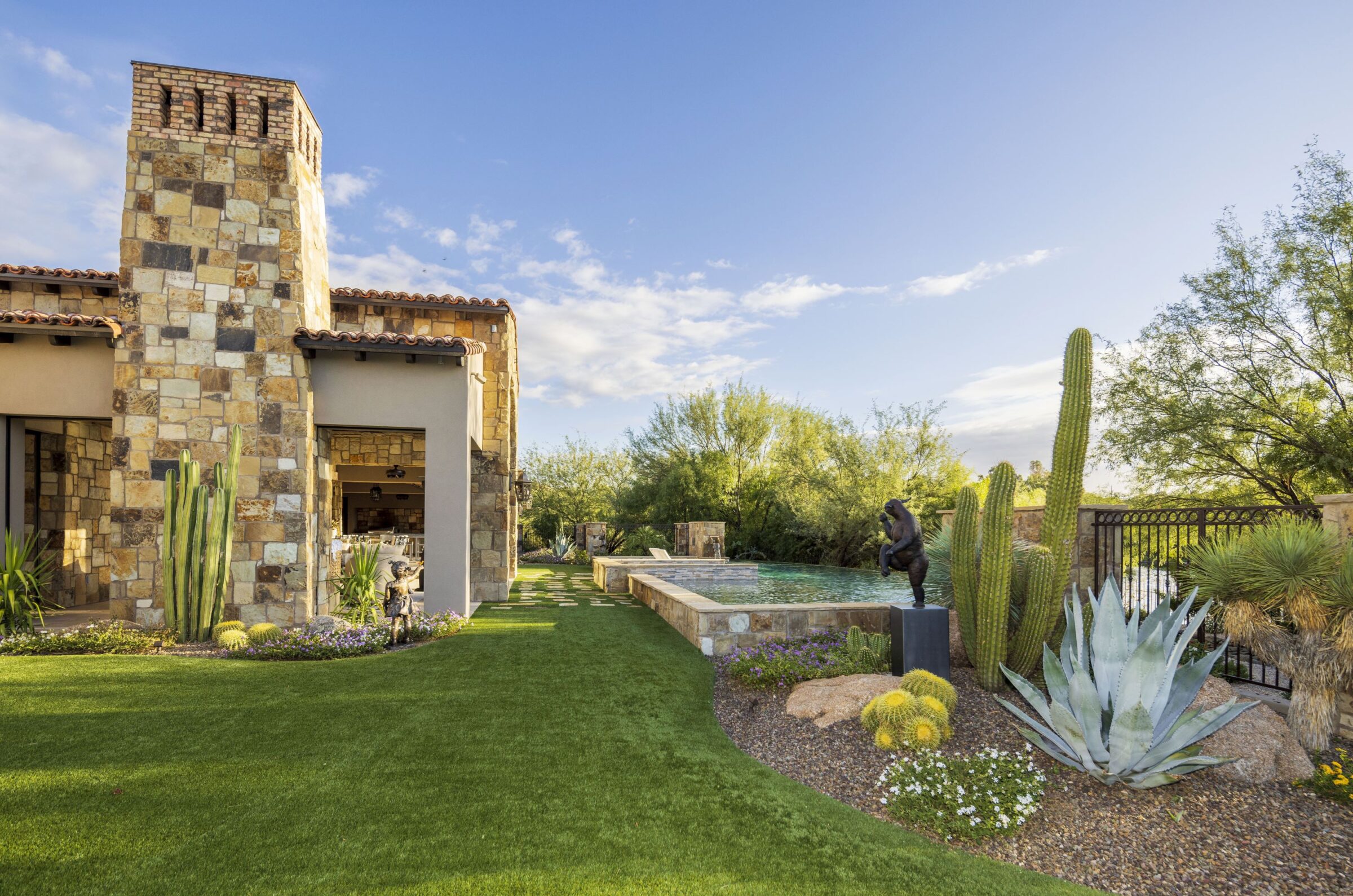 Stone house with large chimney, landscaped yard featuring desert plants, and a swimming pool. A statue is near cacti and agave.