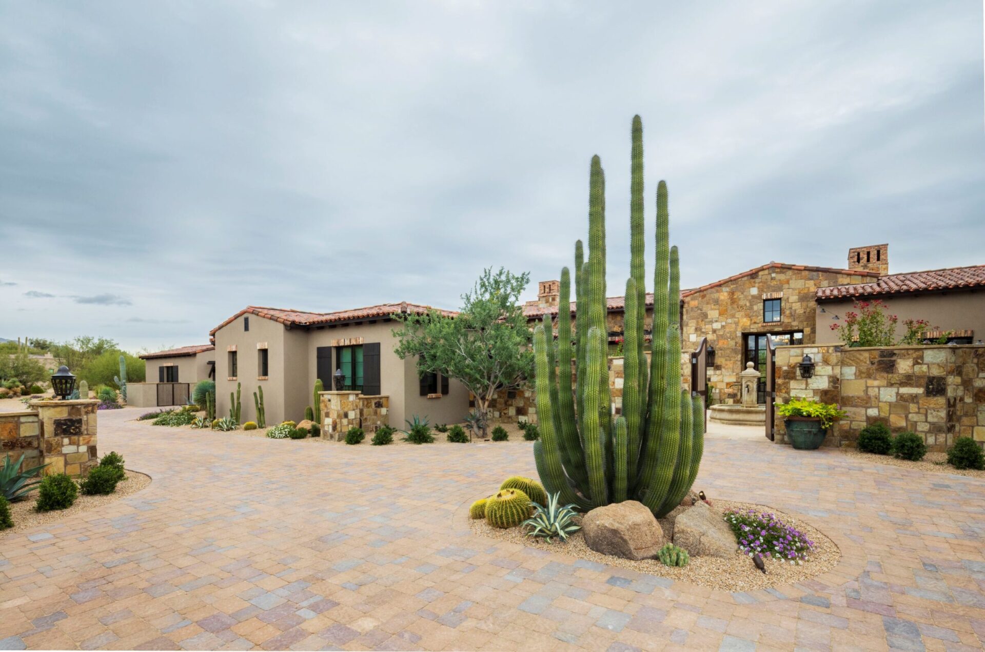 A desert-styled house with a stone facade and red roof, surrounded by cacti and plants, set against a cloudy sky.