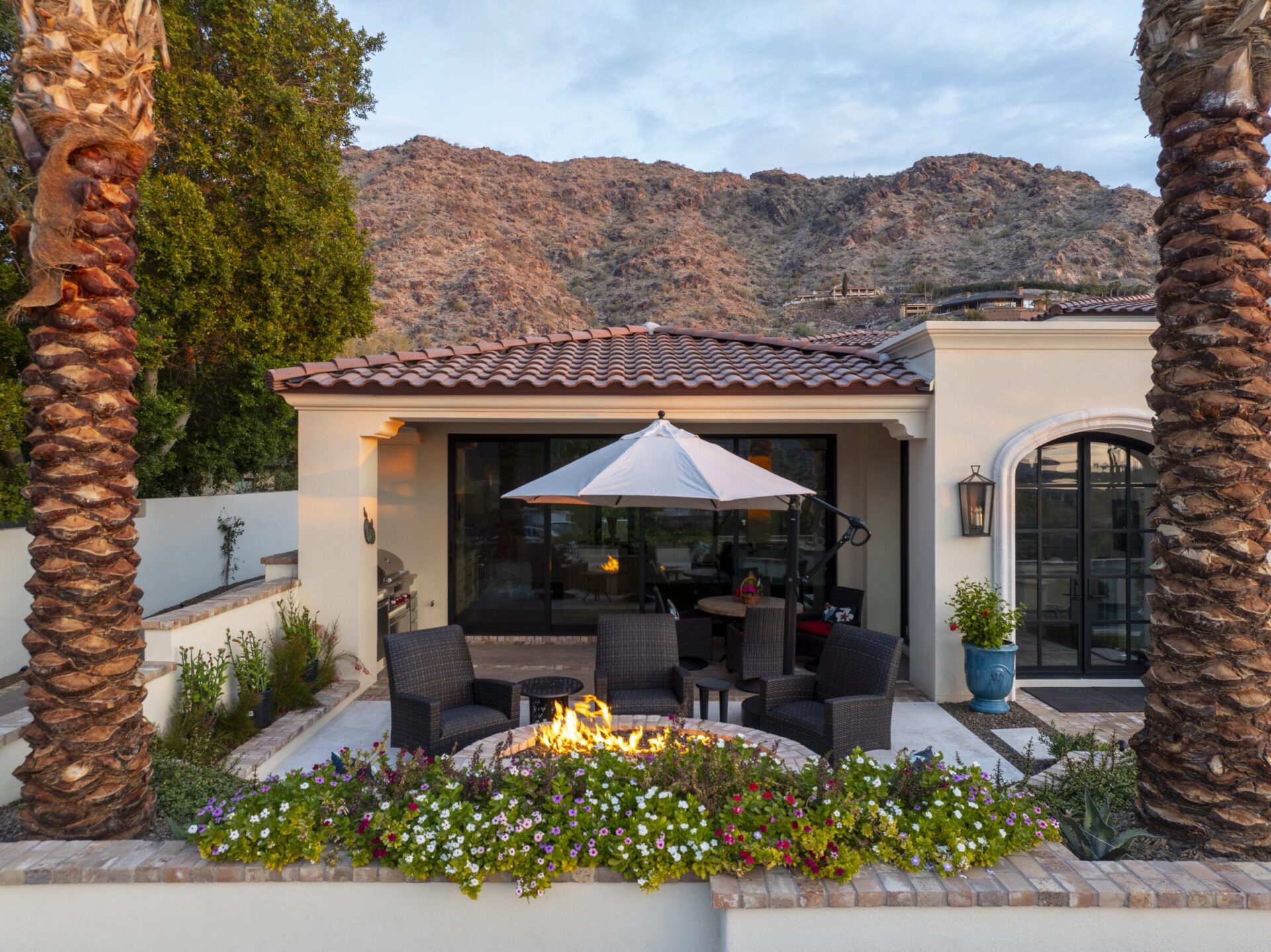 Patio with fire pit, wicker chairs, and umbrella. Surrounded by colorful flowers and palm trees. Set against rocky hillside backdrop. Cozy outdoor space.