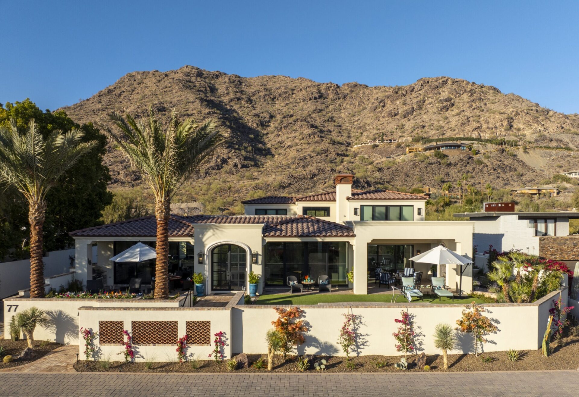 Modern house with palm trees situated in front of a desert mountain backdrop. Outdoor seating with umbrella visible on the patio.