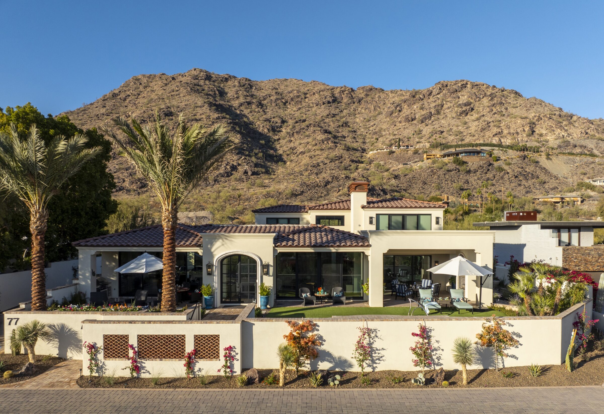 Modern house with a tiled roof, palm trees, and landscaped garden, set against a mountainous backdrop under a clear blue sky.