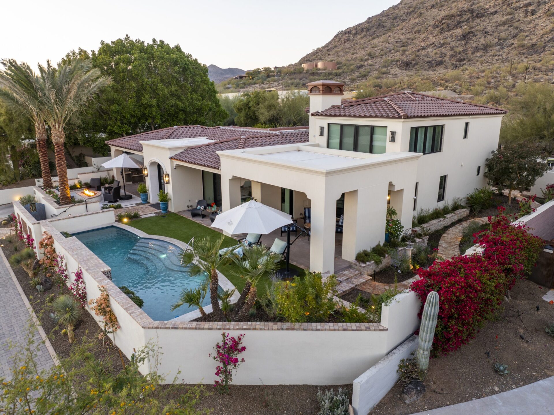 Modern villa with a pool, palm trees, and umbrellas in a mountainous desert landscape, surrounded by lush greenery and flowering plants.