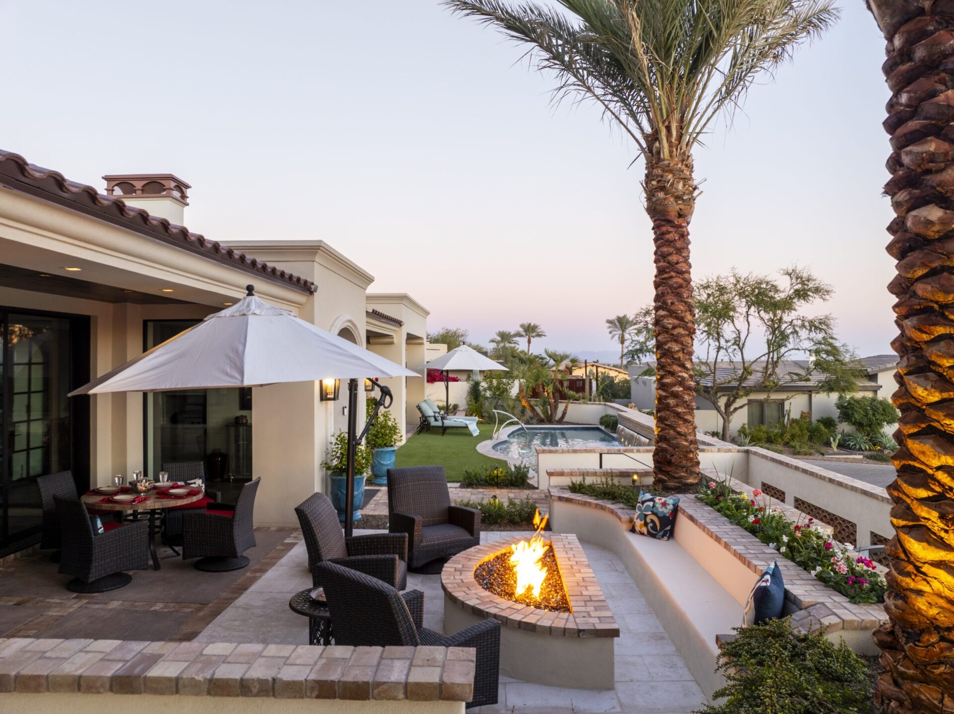 Luxurious patio with fire pit, wicker furniture, palm trees, swimming pool, and outdoor seating. Sunset ambiance creates a warm and inviting atmosphere.