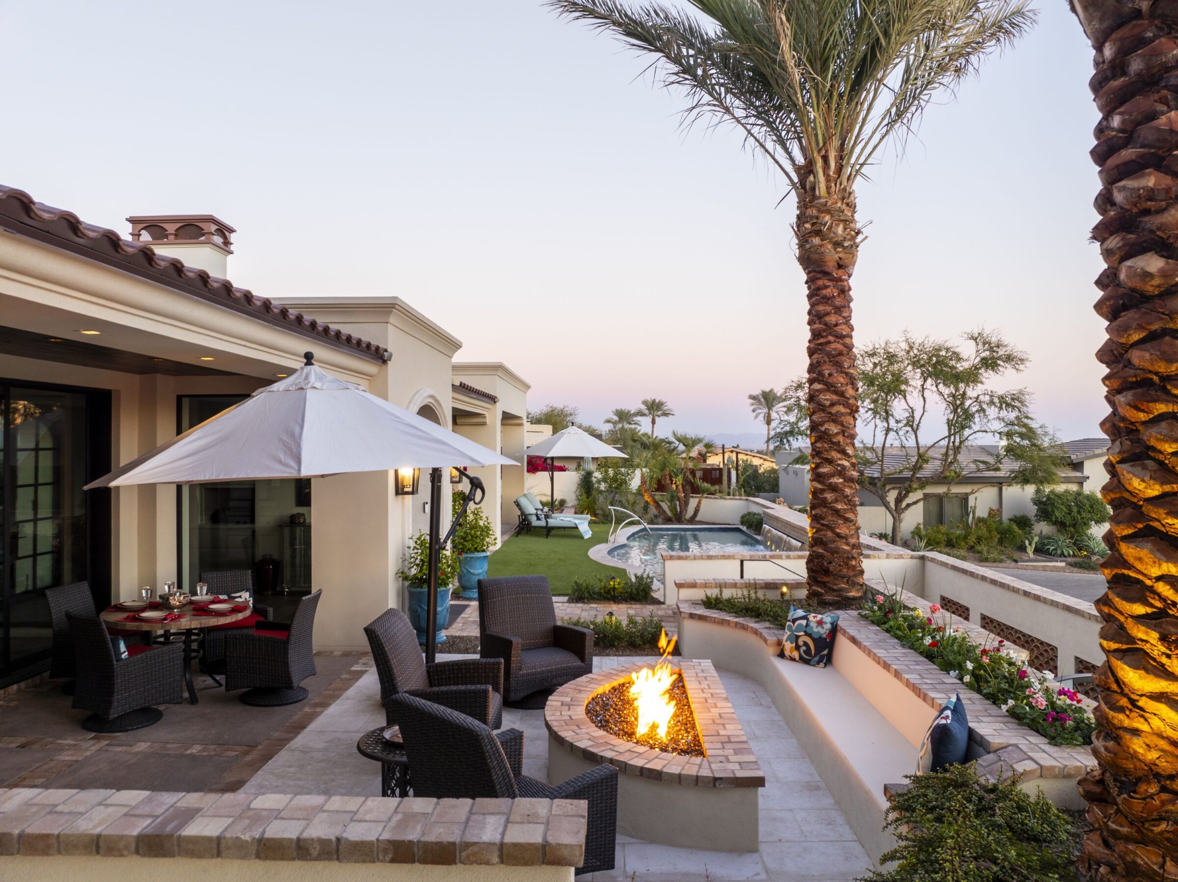 Luxurious backyard with a fire pit, patio furniture, pool, umbrellas, and palm trees, creating a serene and inviting outdoor living space.