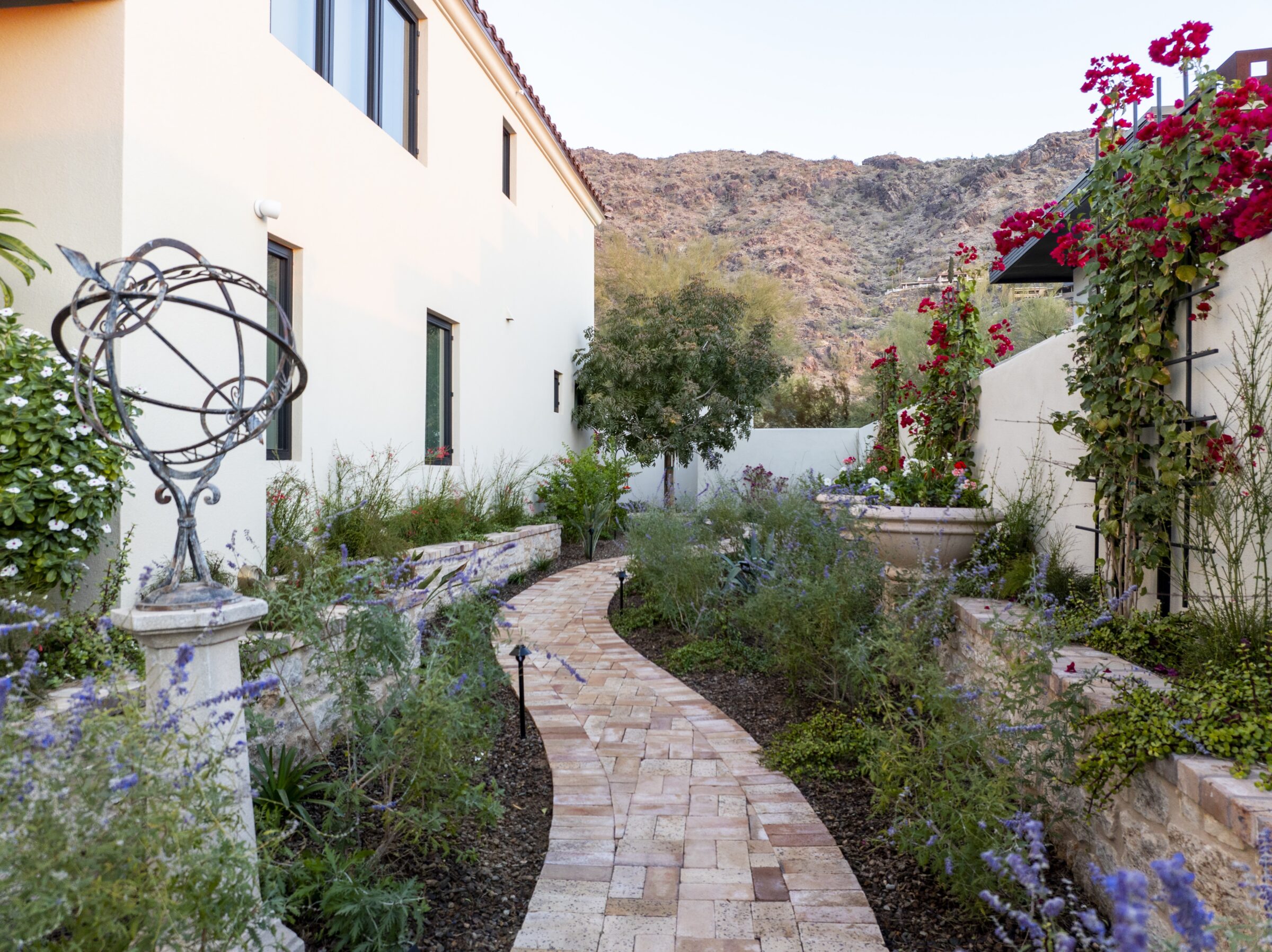 A narrow brick path winds through a lush garden with flowers, leading to mountains under a clear sky, beside beige house walls.
