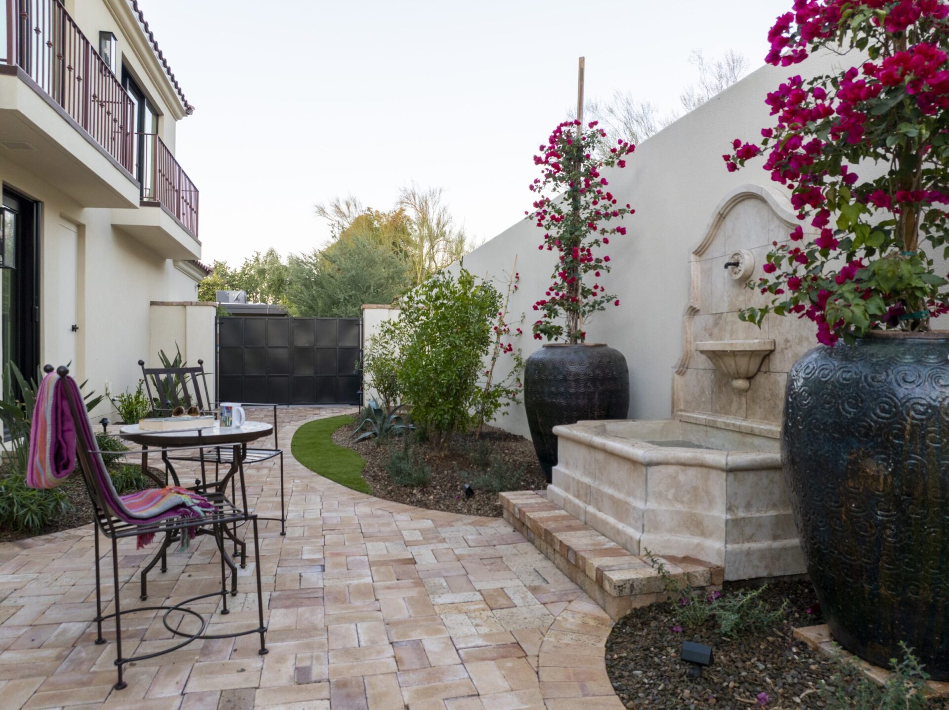 A serene courtyard with a stone fountain, potted flowers, patio table, and chairs. Clean pathways and greenery enhance the peaceful atmosphere.