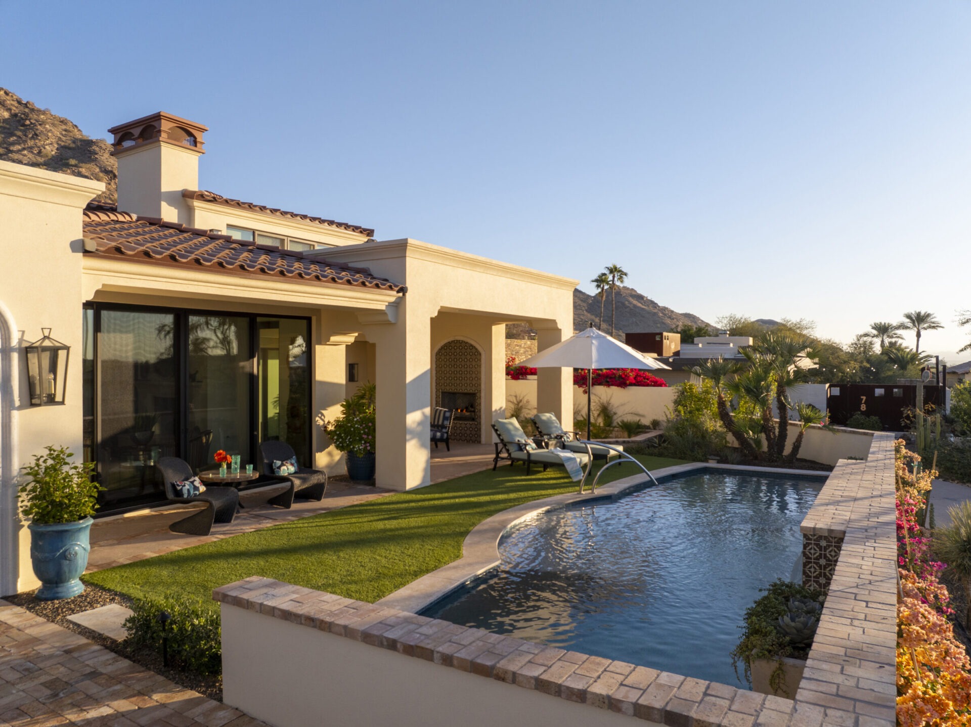 A modern house with a pool, patio, lounge chairs, umbrella, mountains in the background, surrounded by palm trees and colorful garden.