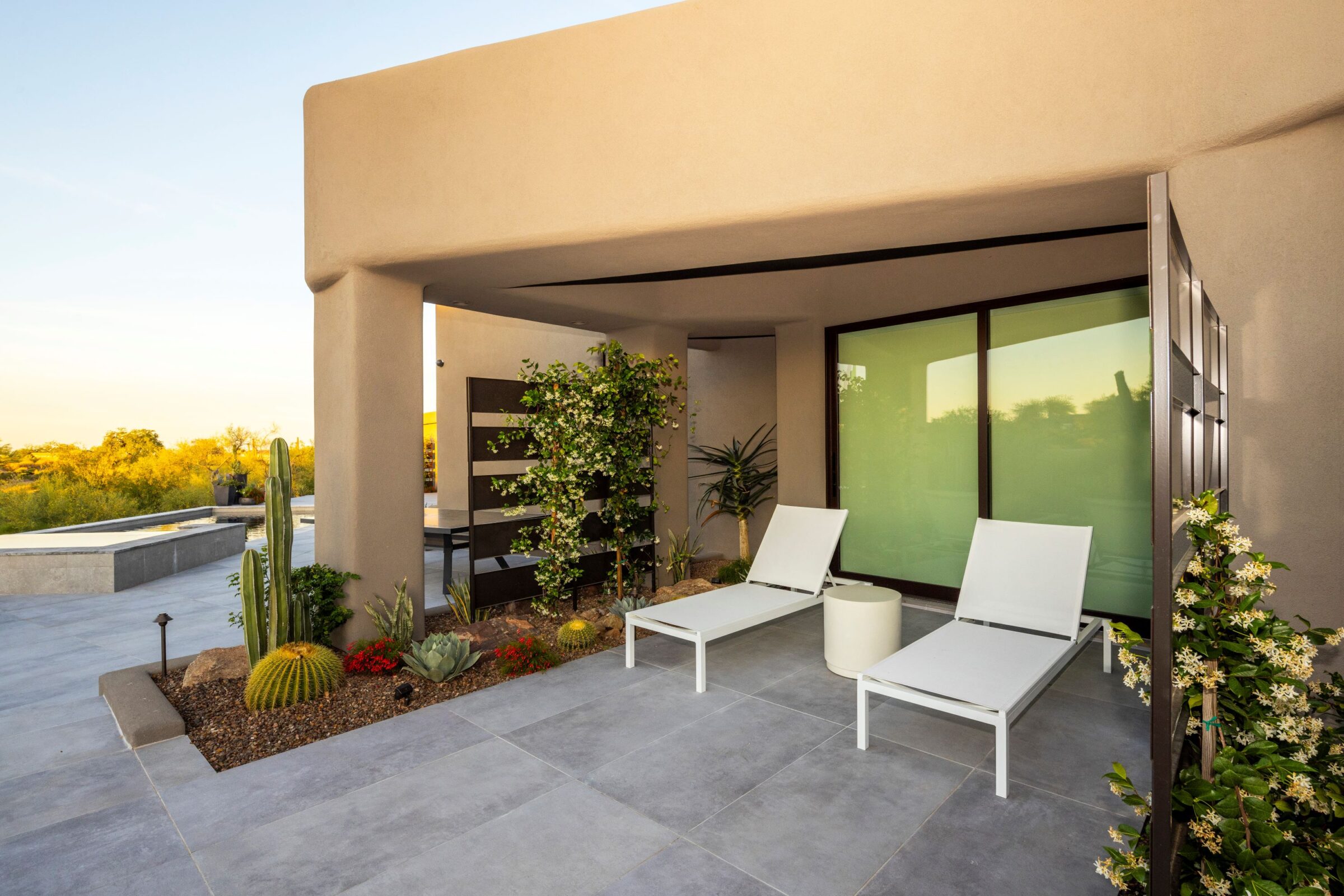 Modern patio featuring two white loungers, cacti, and a small garden, with a contemporary architectural style and natural landscape in the background.