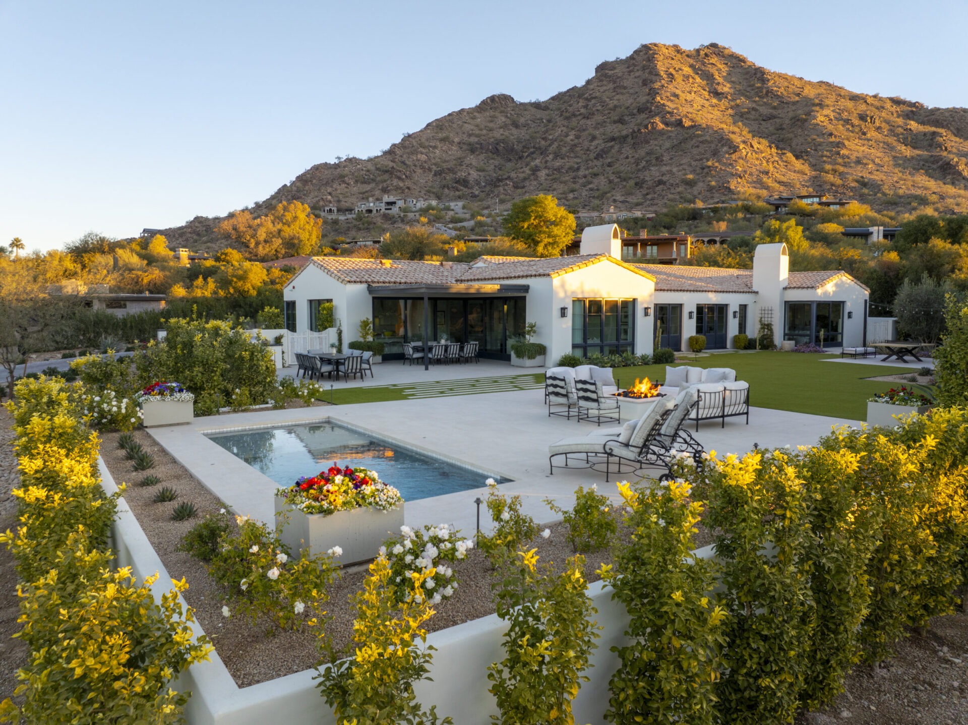 A modern villa with a pool and patio seating sits against a mountainous backdrop, surrounded by lush greenery and flowers under a clear sky.