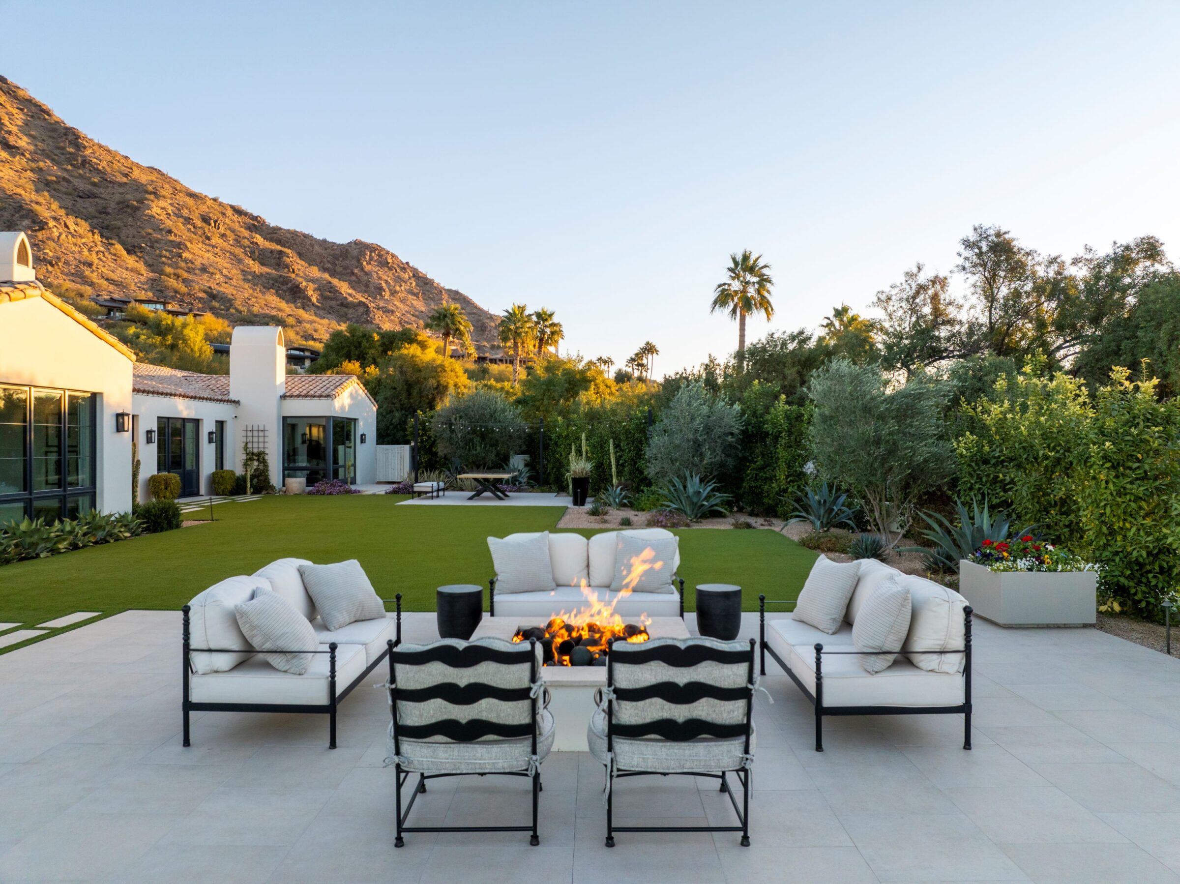 Outdoor seating area with fire pit, surrounded by lush greenery, facing a scenic mountain backdrop. Comfortable sofas and chairs create a relaxing ambiance.