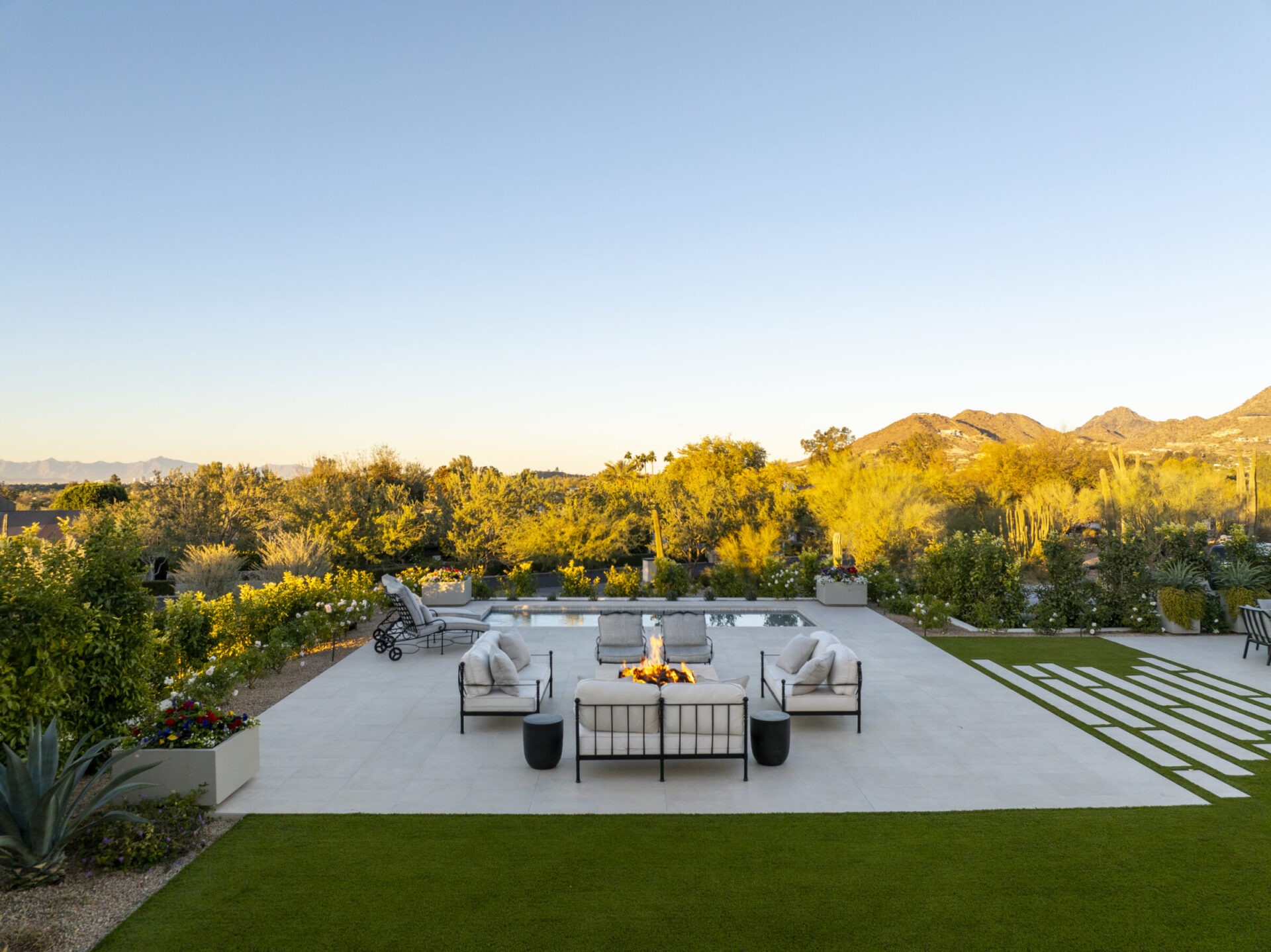 Outdoor patio with modern seating around a fire pit, lush greenery, and distant mountain views under a clear blue sky at sunset.