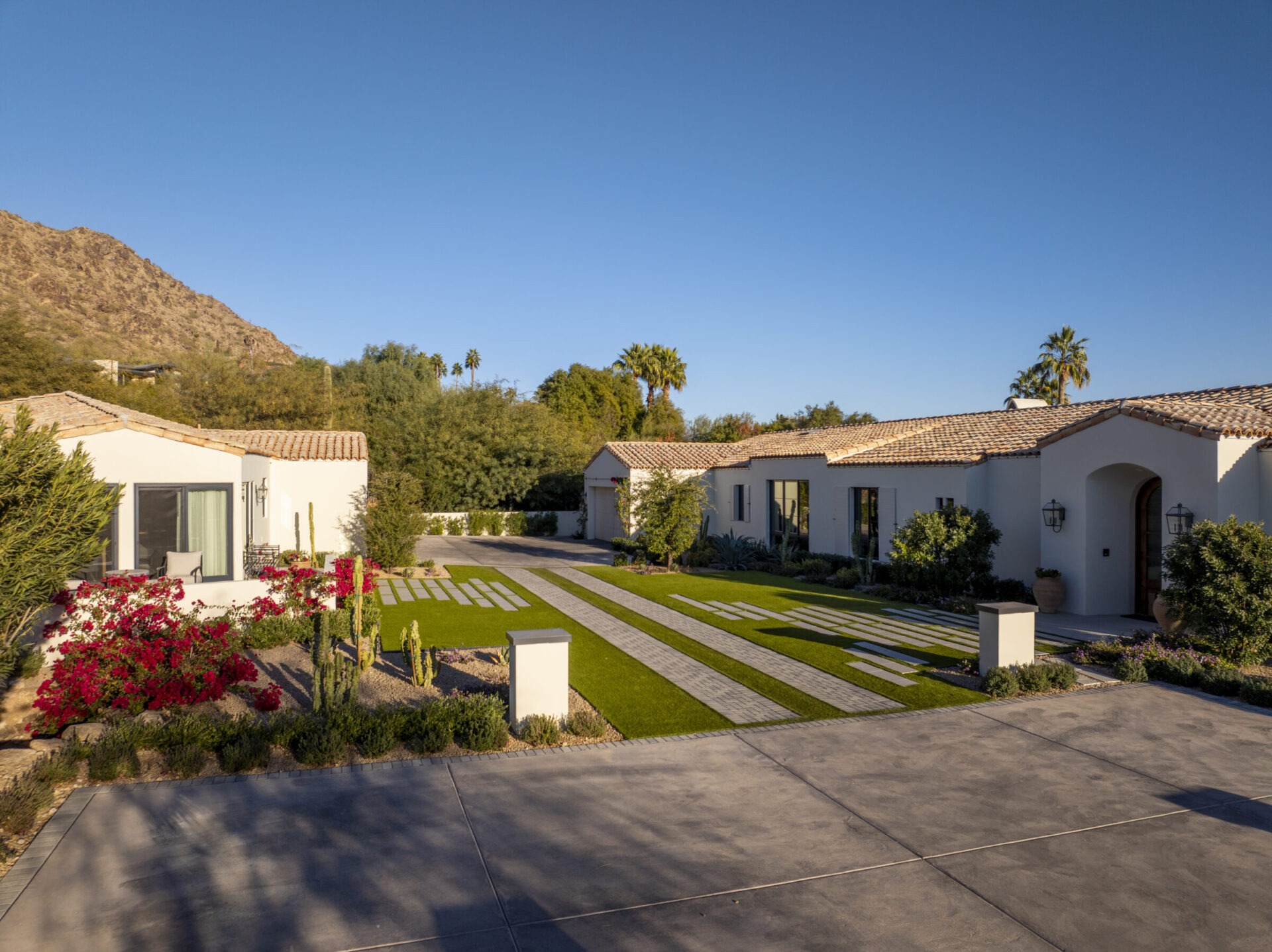 Spacious modern house with tiled roof, lush green lawn, flowering plants, and trees, set against a backdrop of mountains and a clear sky.