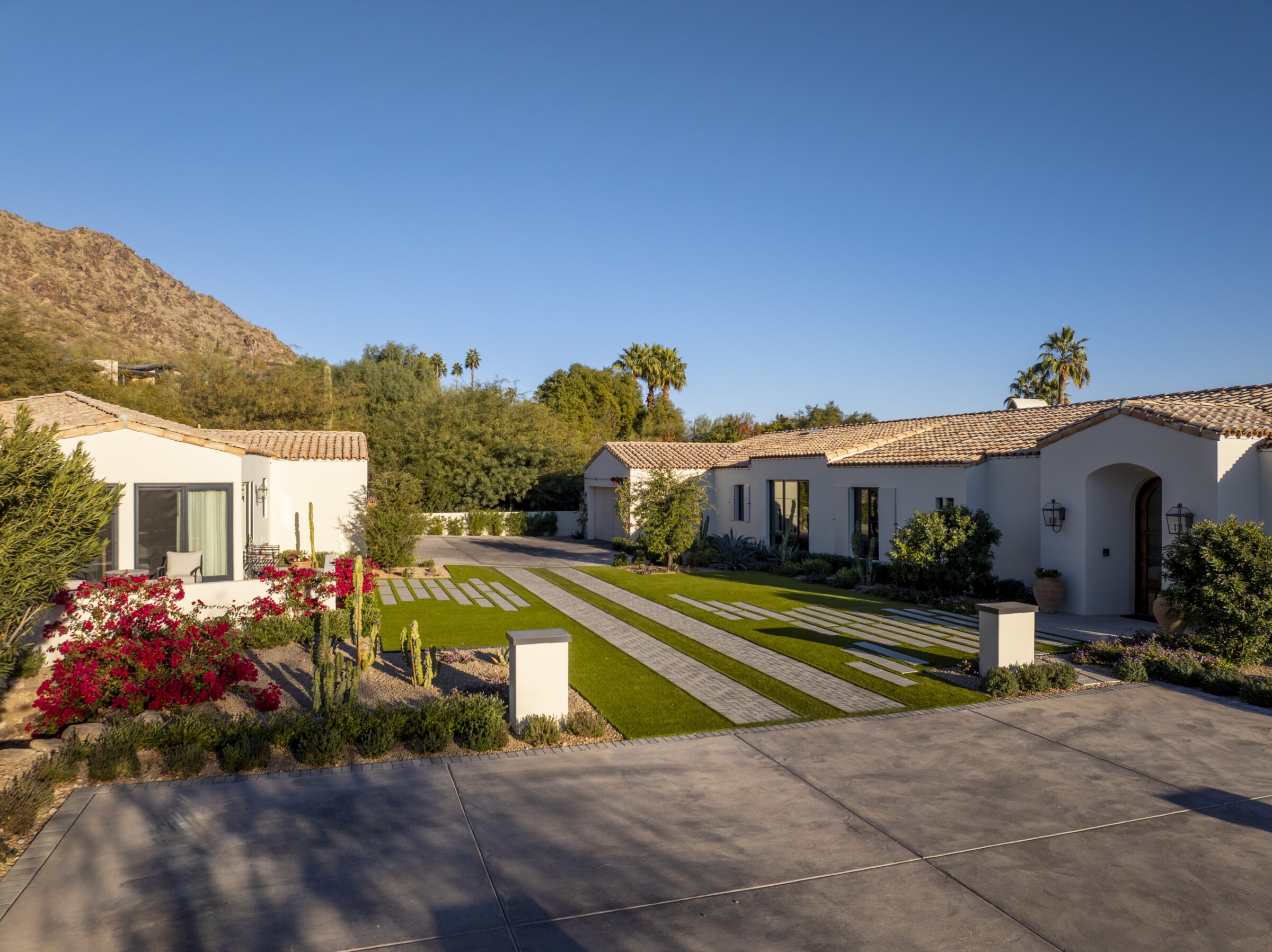 Spacious modern house with tiled roof, lush green lawn, flowering plants, and trees, set against a backdrop of mountains and a clear sky.