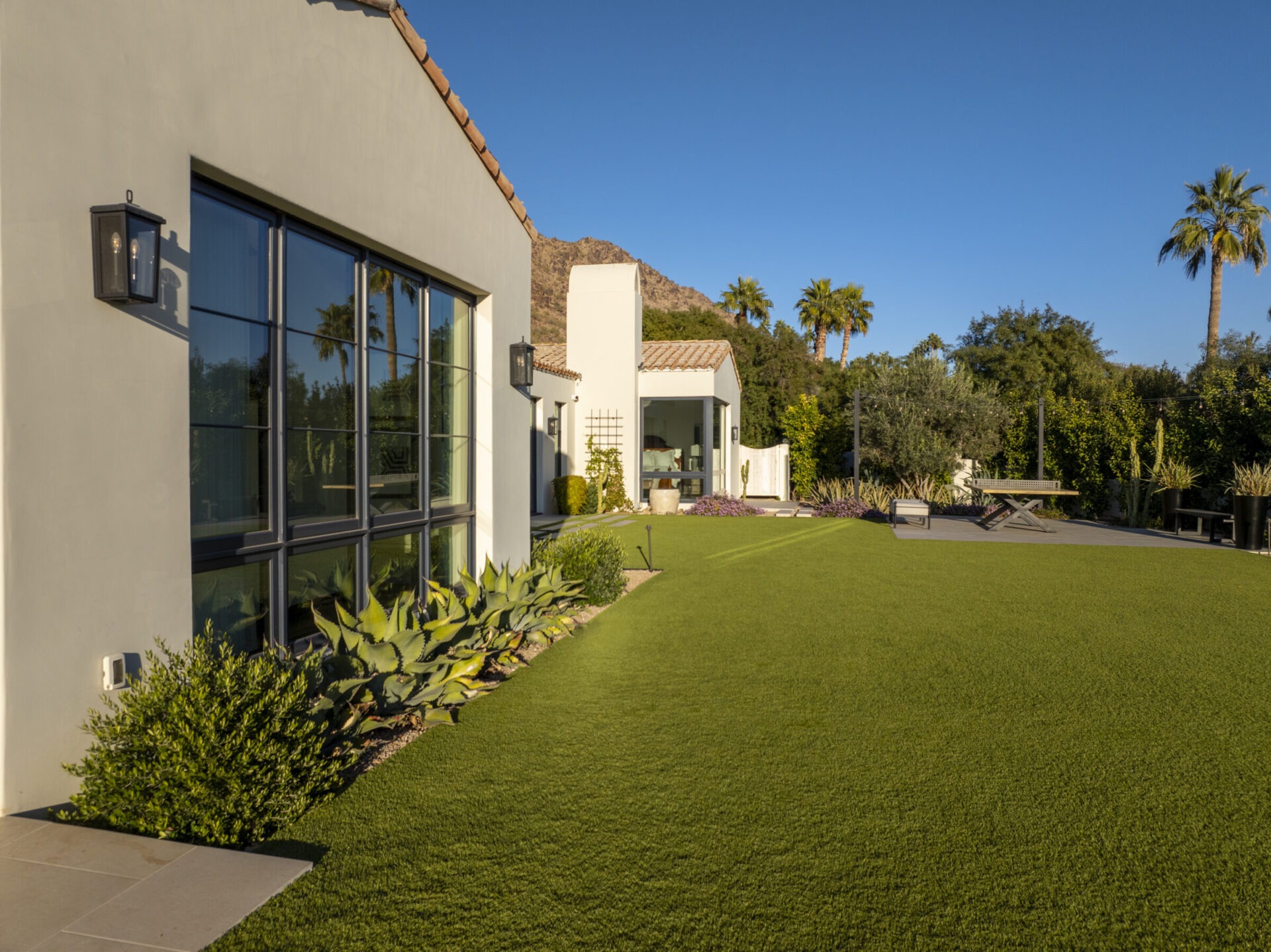 Modern house with large windows, surrounded by lush greenery and palm trees. The lawn is expansive, and mountains are visible in the background.