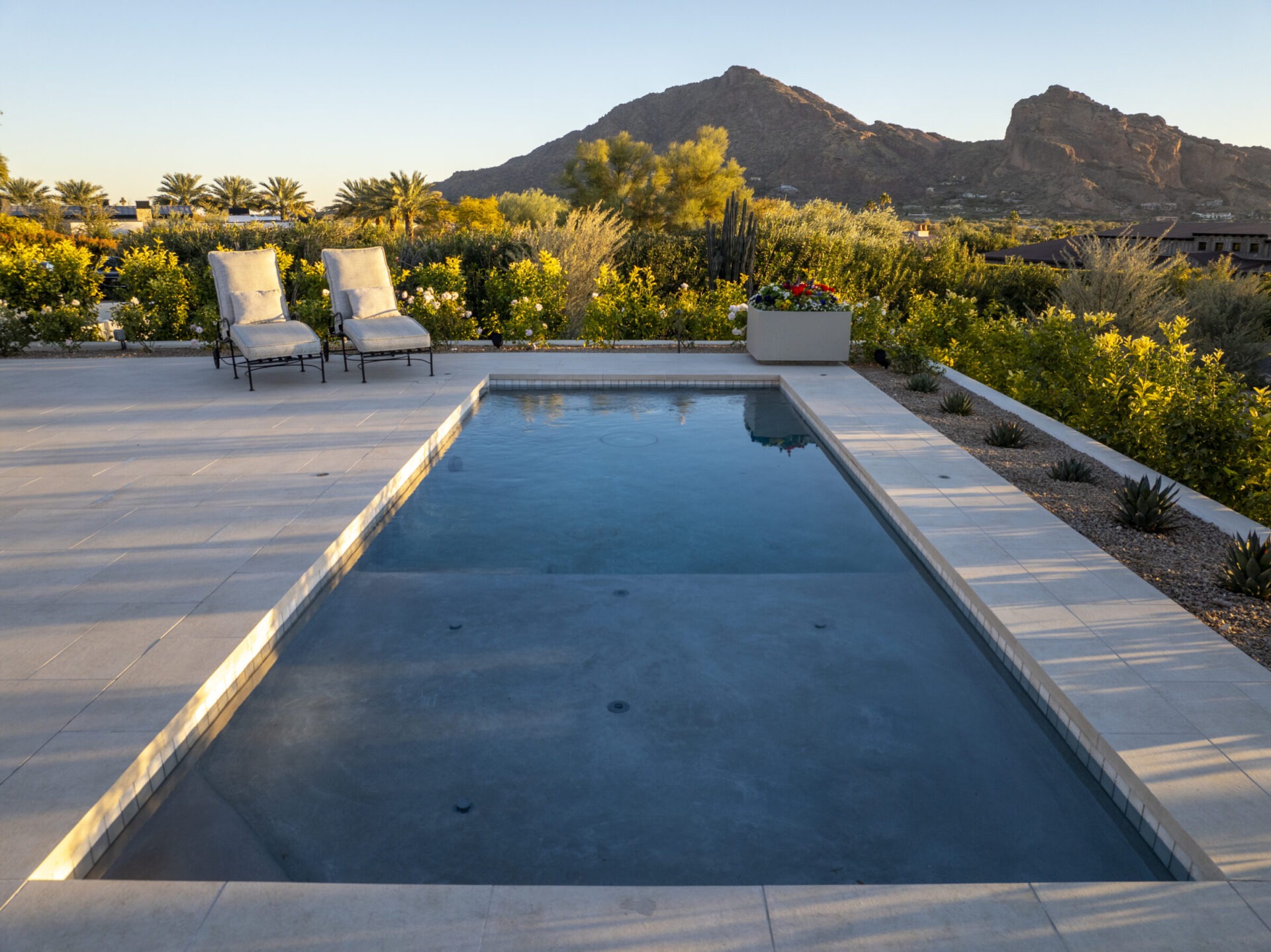 A serene poolside patio with two lounge chairs, surrounded by lush greenery, overlooks majestic mountain terrain under a clear sky.