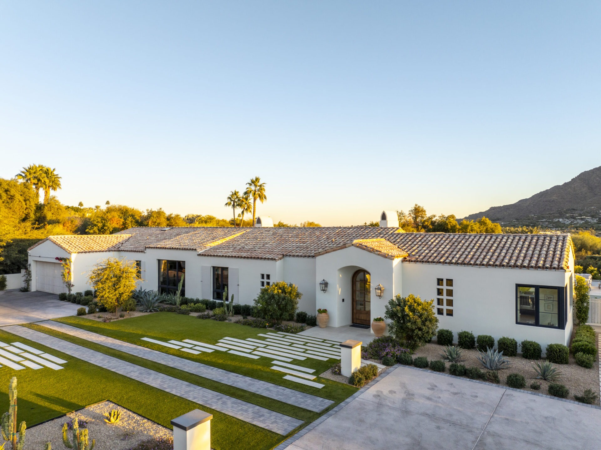 Single-story white house with terracotta roof, landscaped garden, palm trees, and mountain backdrop under a clear blue sky. Modern design elements are visible.