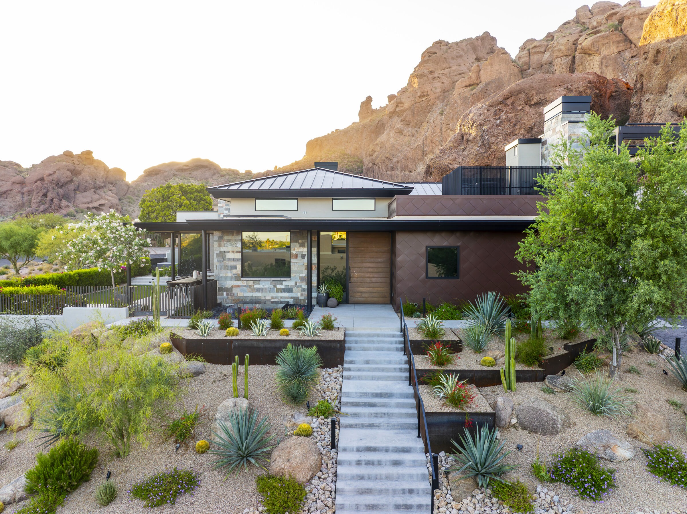 Modern house with desert landscaping and stone facade, set against rocky cliffs. Features a staircase, numerous cacti, and lush greenery.