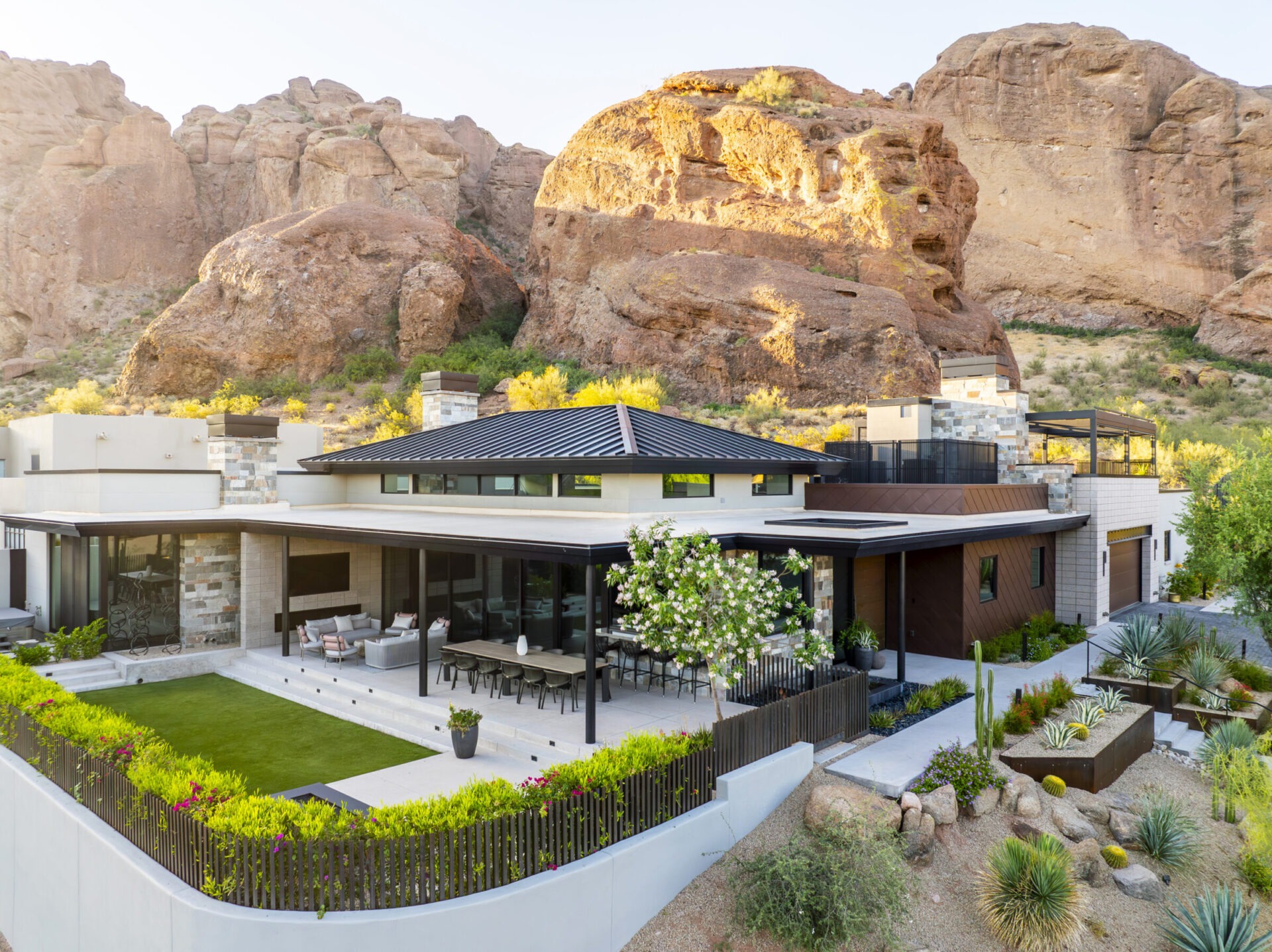 Modern house with large patio set among stunning, rocky desert cliffs. Lush greenery surrounds the home, blending luxury with natural beauty.