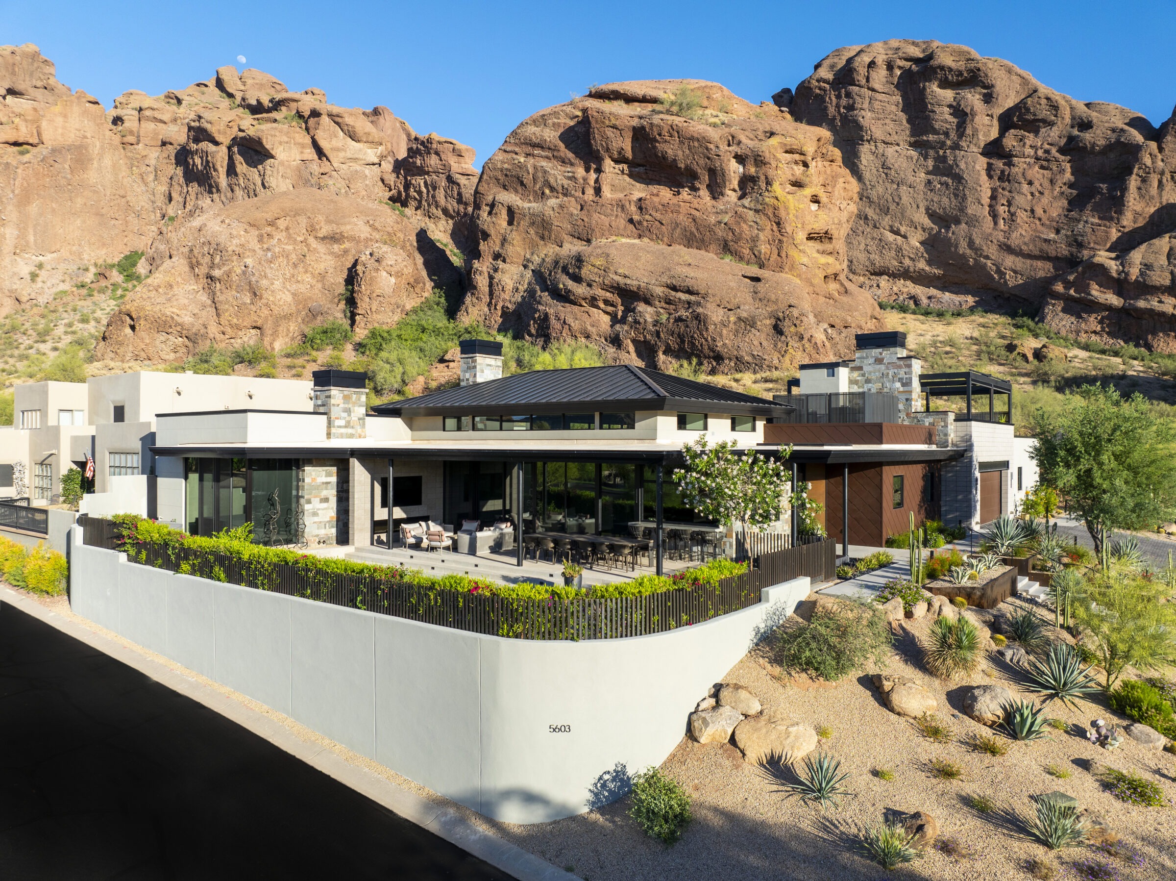 A modern house nestled against rocky cliffs, surrounded by desert landscaping, under clear blue skies. The area appears serene and secluded.