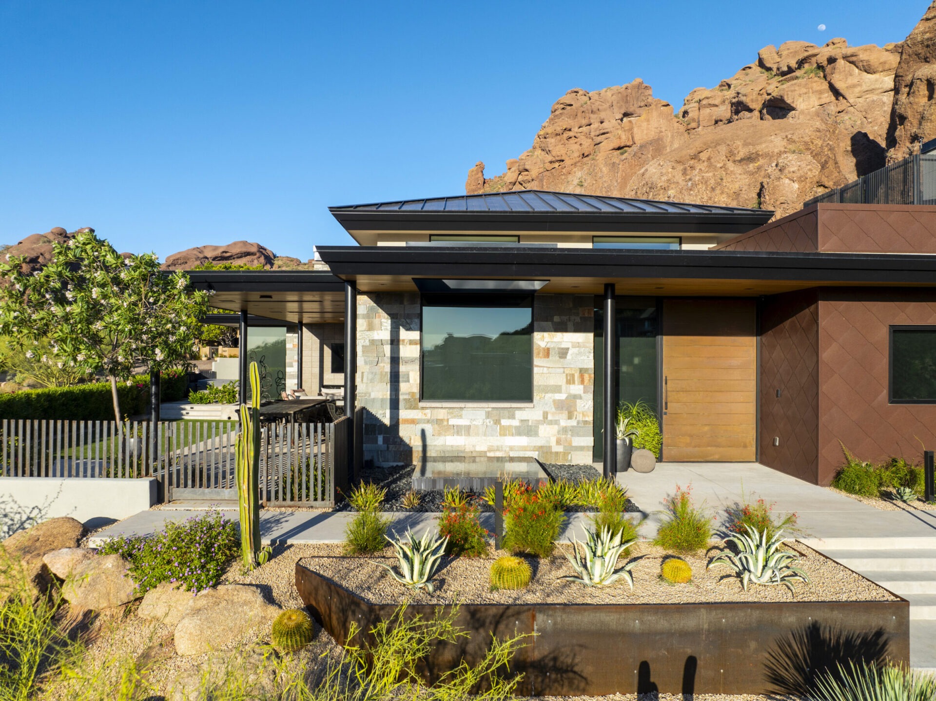 Modern house with desert landscaping and cacti, set against rocky mountain backdrop under a clear blue sky. No people are visible.