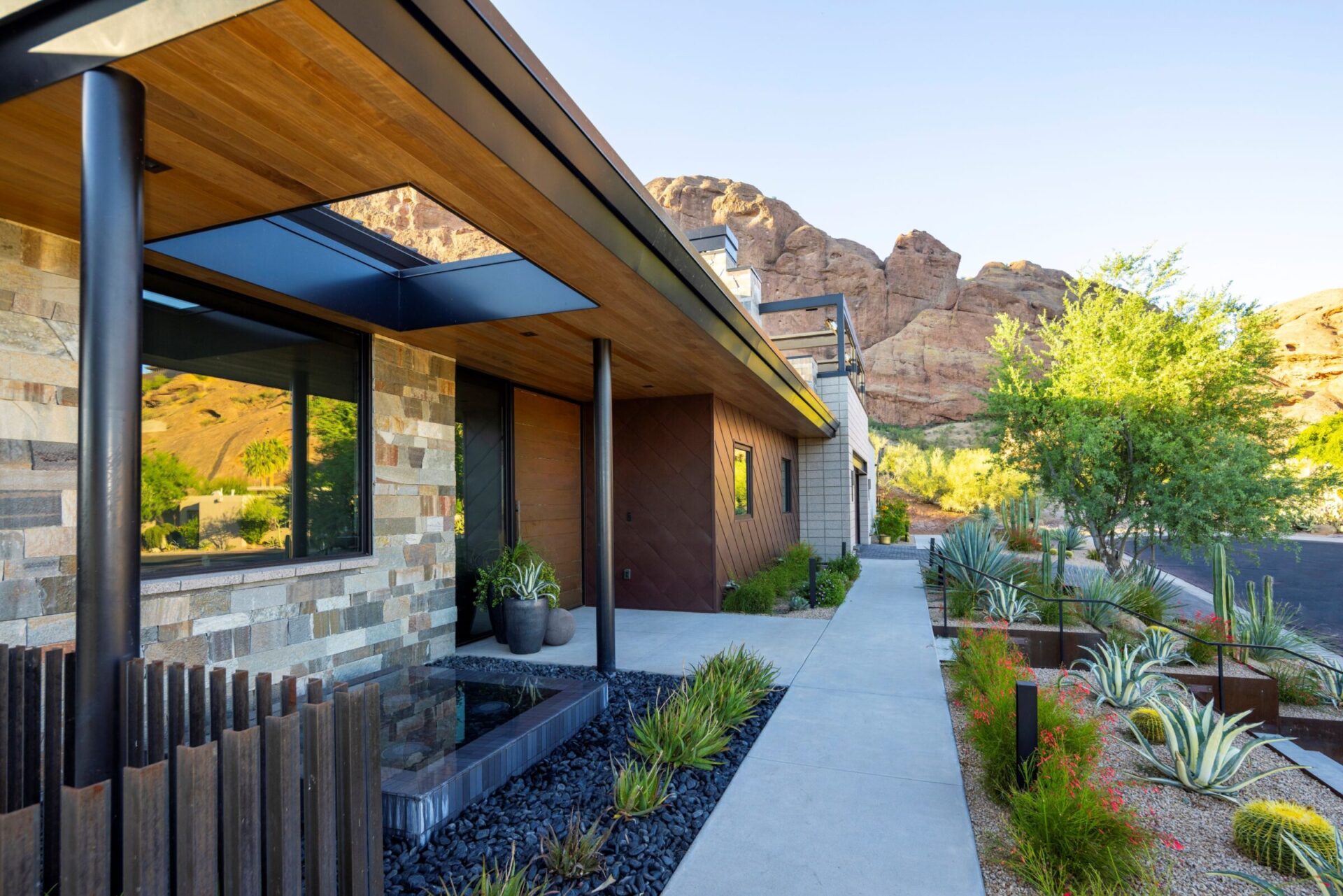 Modern house with stone facade and desert landscaping, set against a backdrop of rocky mountains under clear blue sky. Pathway leads alongside agave plants.