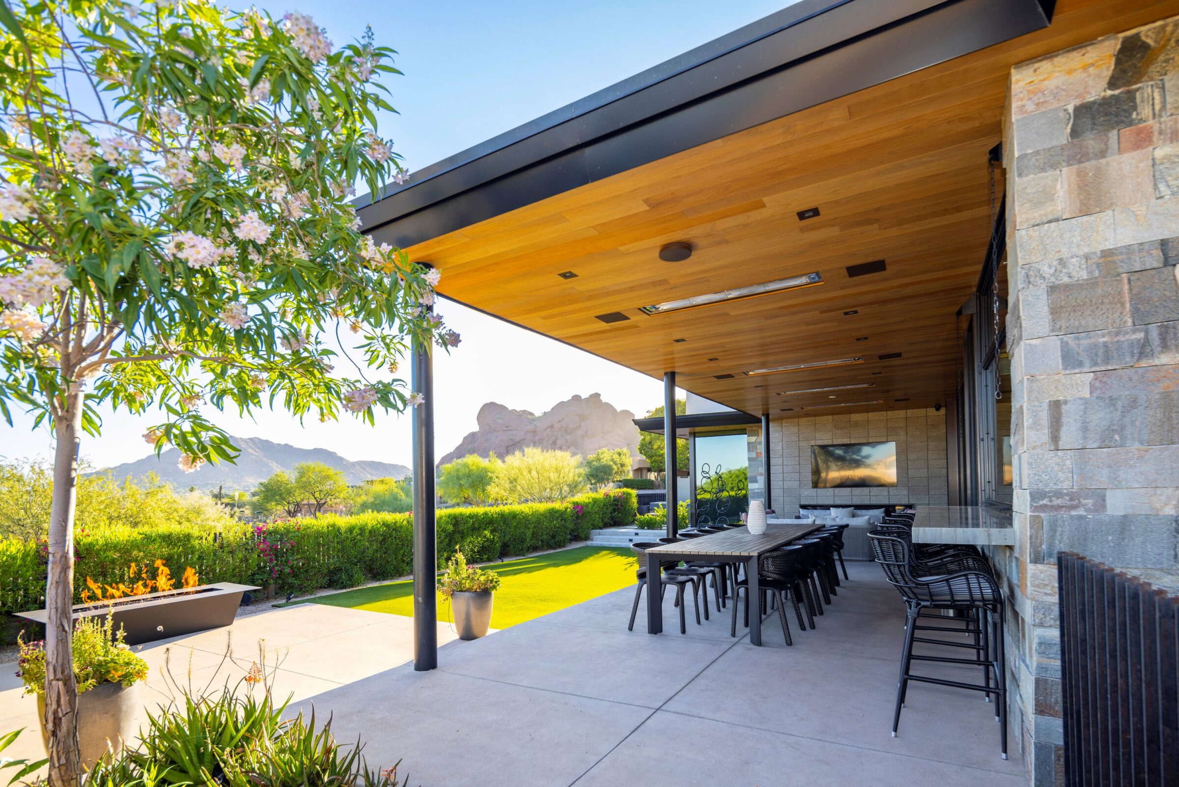 Modern patio with a wooden ceiling, fire pit, and outdoor seating area. Lush greenery and distant desert mountains provide a scenic backdrop.