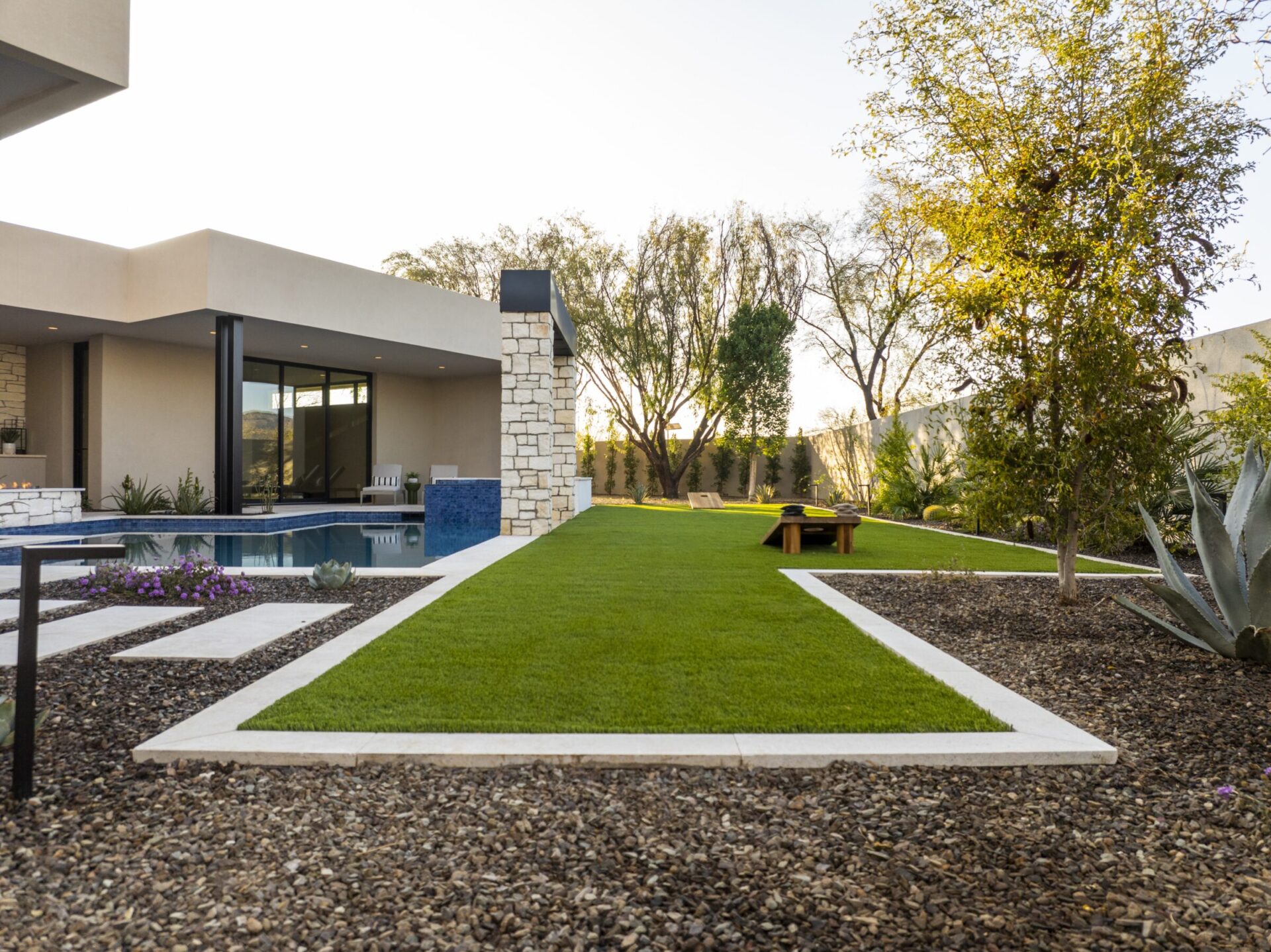 Modern house with a pool and landscaped garden. Stone walls and trees surround a well-maintained lawn under clear skies. No people visible.