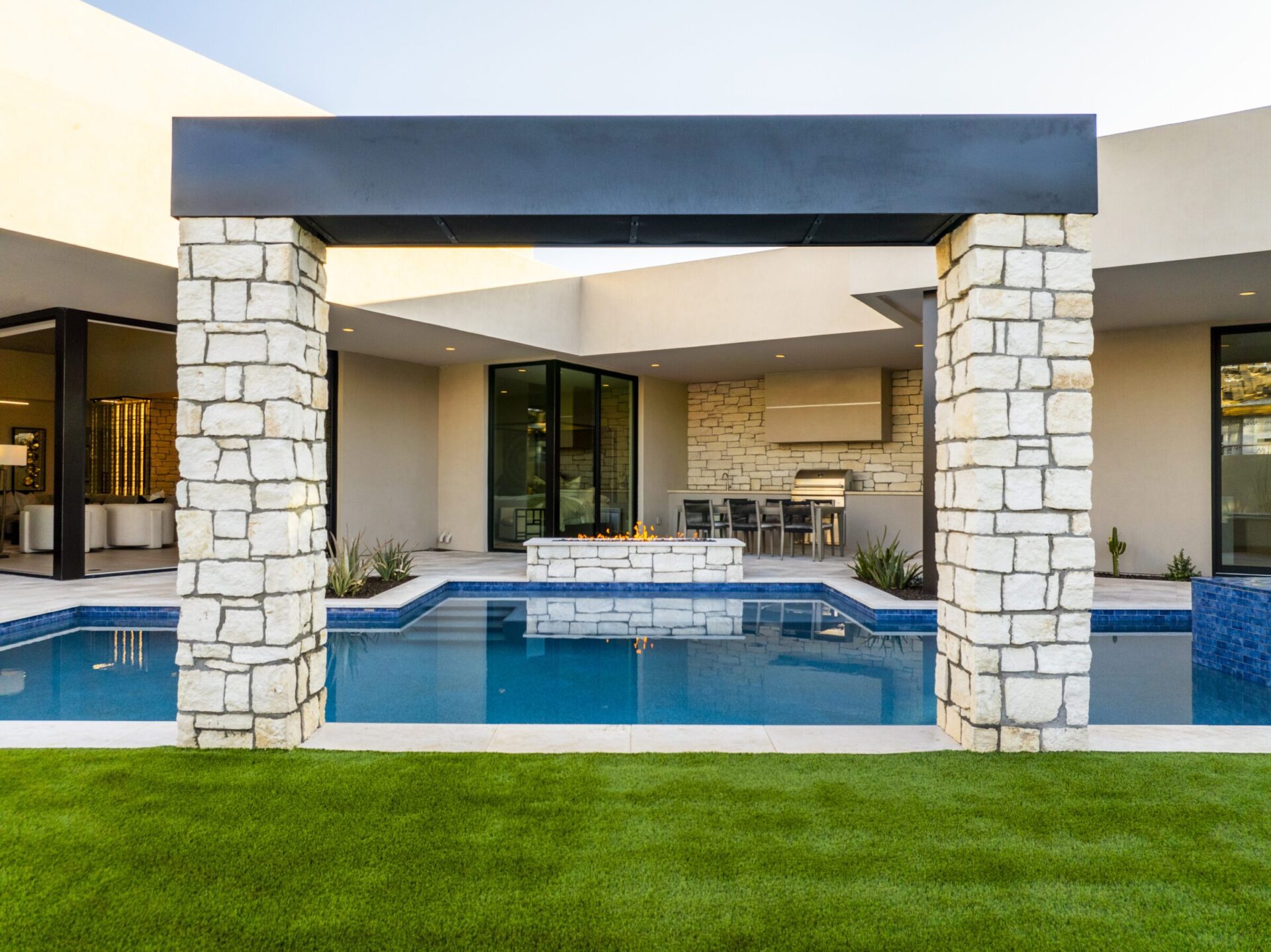 Modern house with stone pillars, pool, and outdoor seating area. Green lawn in foreground, sleek architecture evident throughout the design.